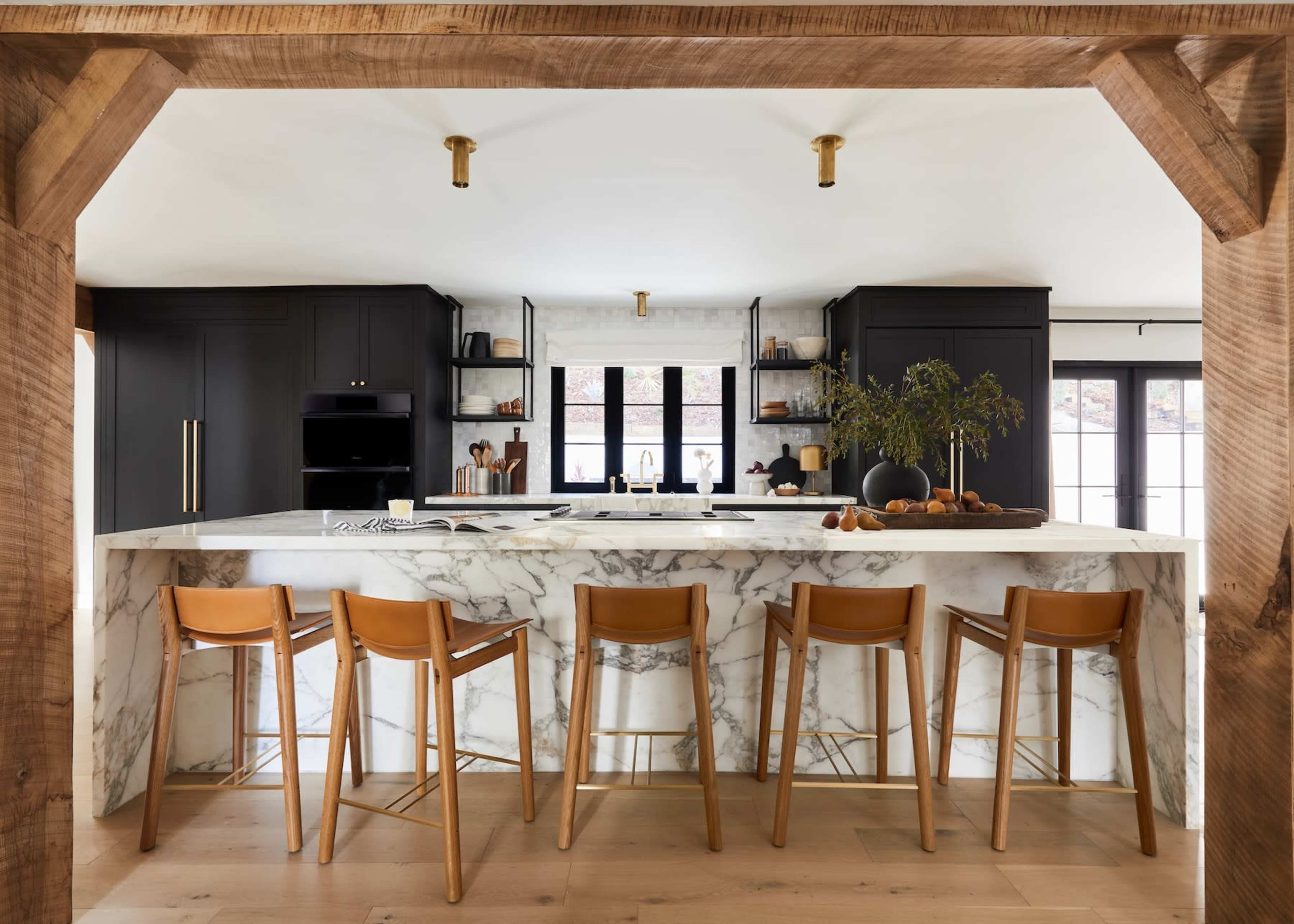 A modern kitchen features a marble island with wooden bar stools and black cabinetry, illuminated by brass fixtures.