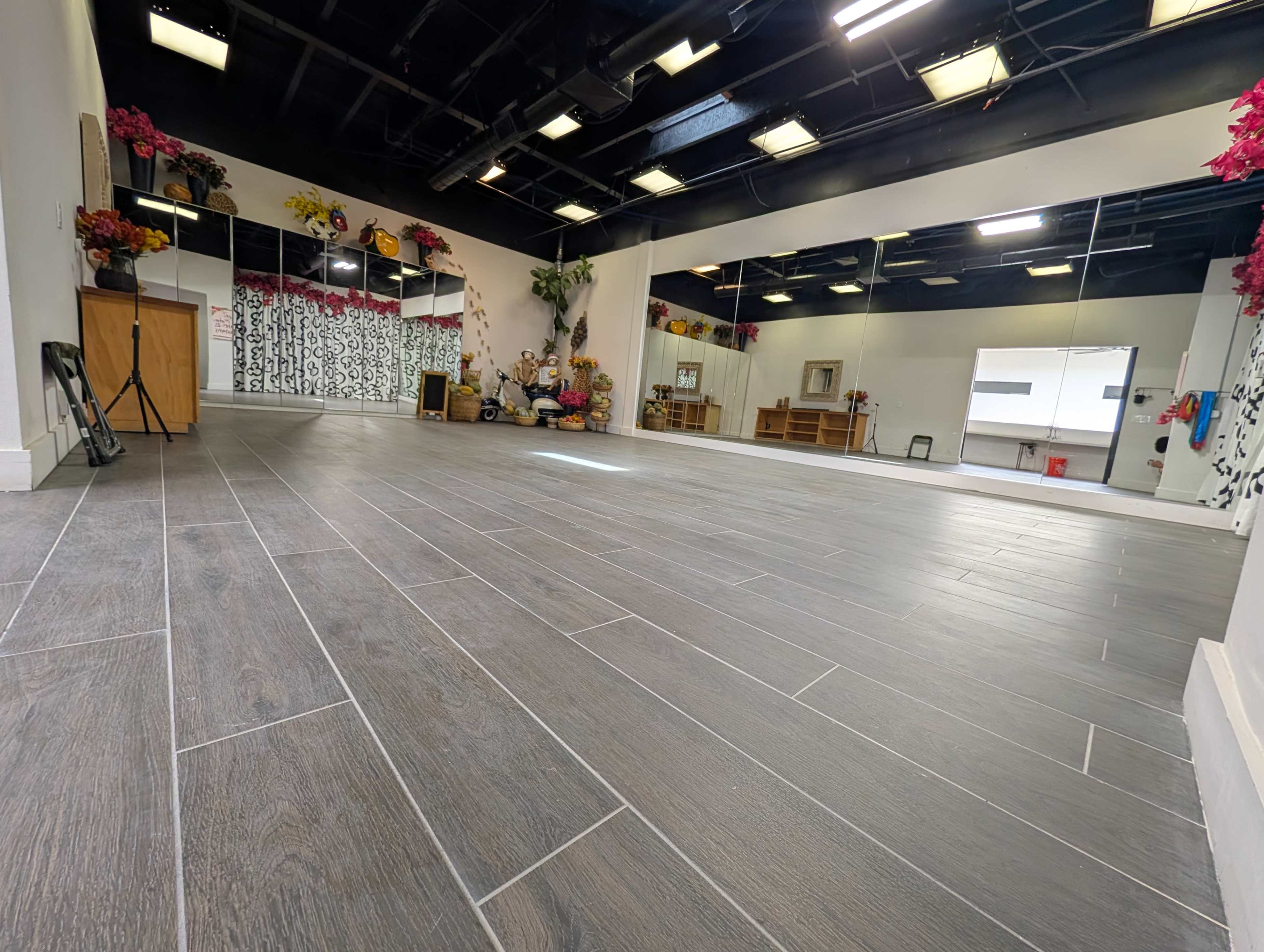 The image shows a spacious, well-lit studio with dark wooden floors, decorated with potted plants and colorful flowers along the walls.