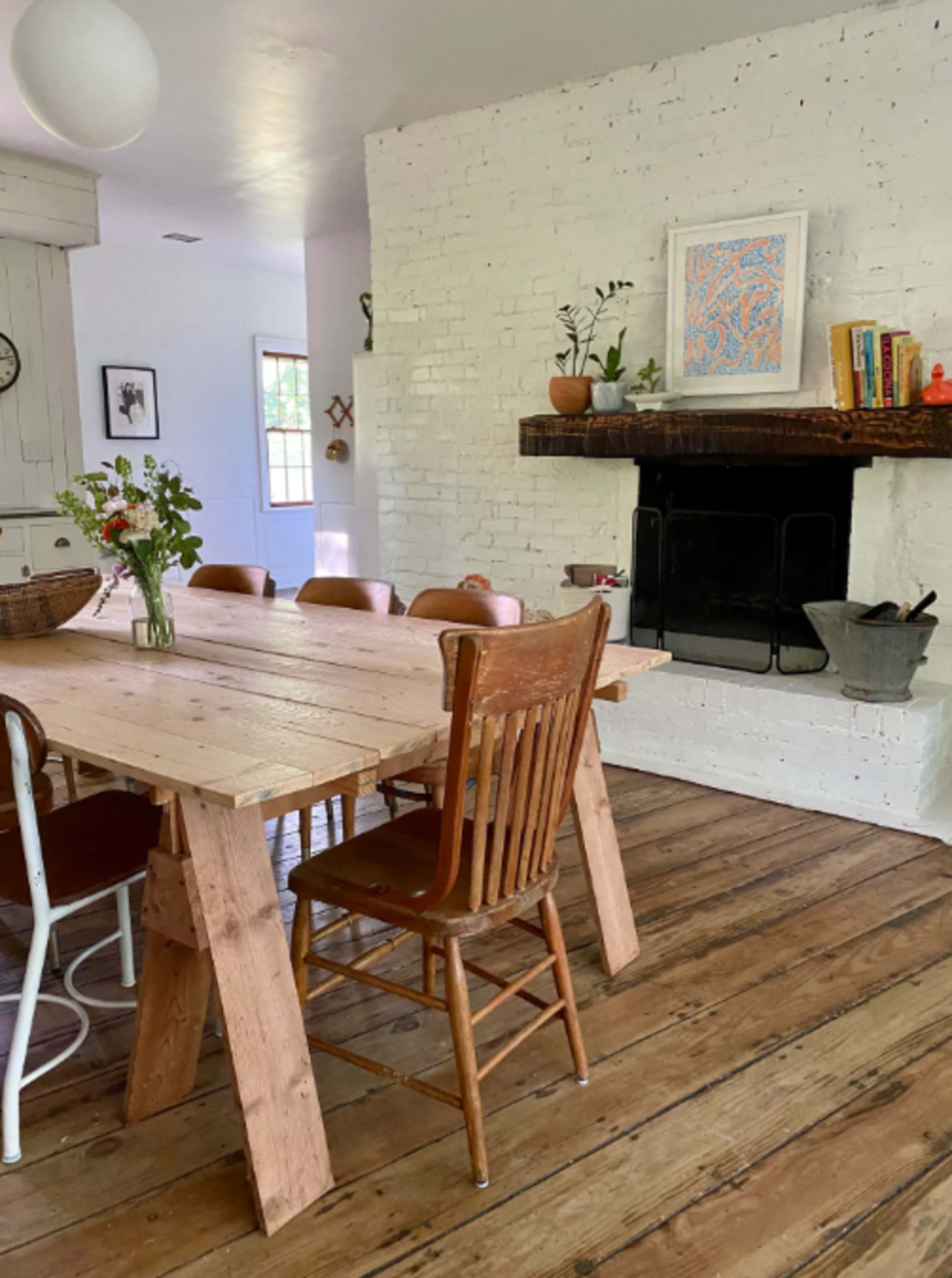 A wooden dining table surrounded by chairs in a bright room with a white brick wall and a fireplace.