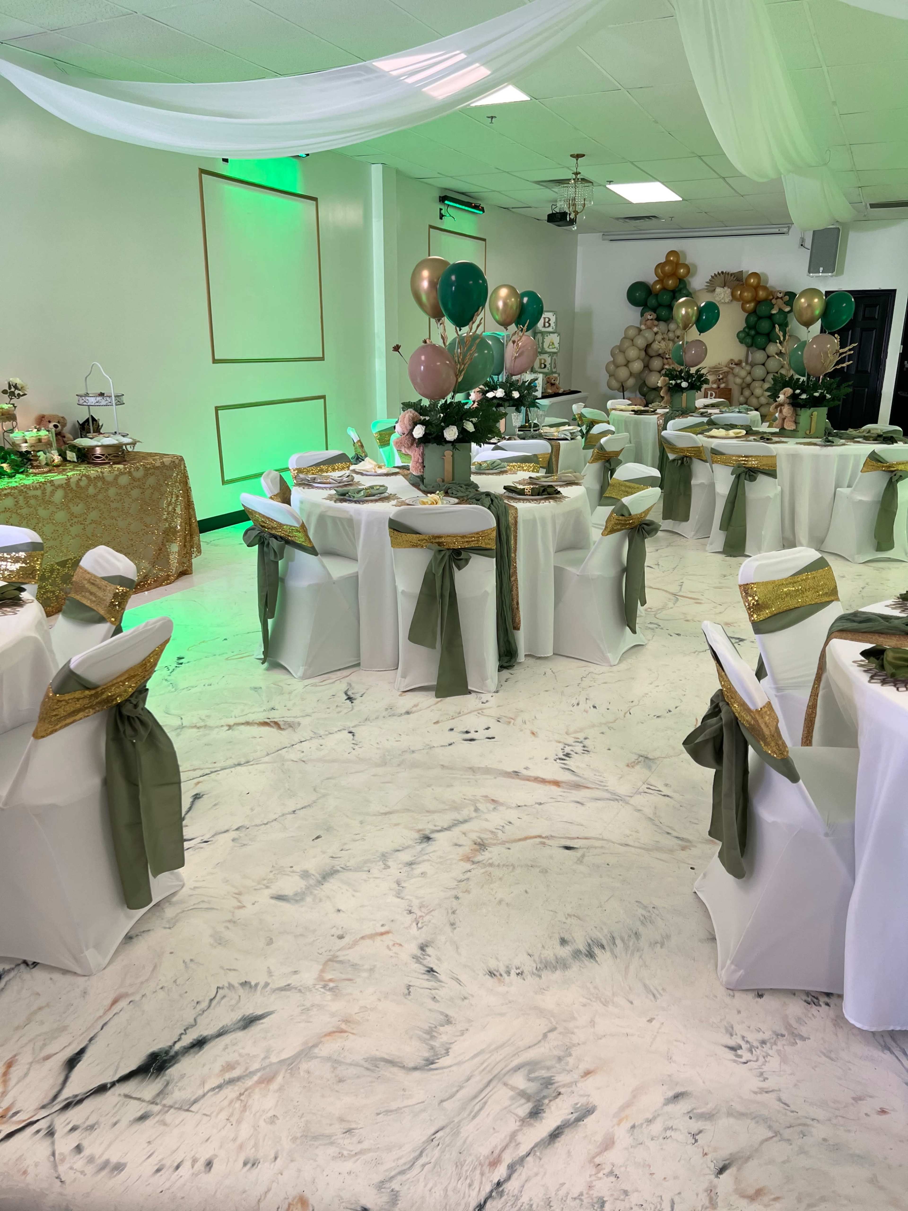 A decorated event space with round tables set for a celebration, featuring white tablecloths, green and gold accents, and various decorative balloons in the background.