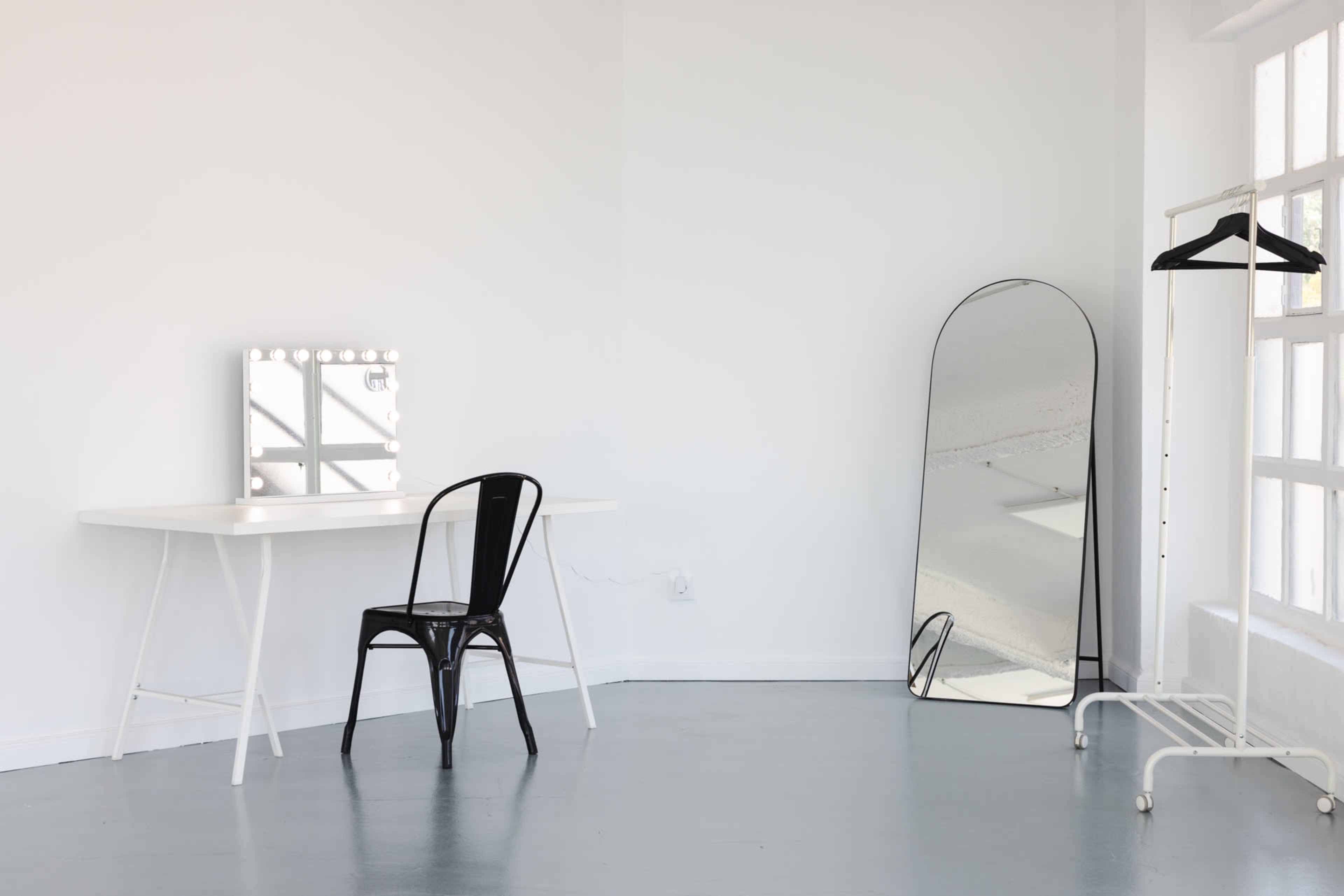 A minimalist room features a white table with a mirror surrounded by light bulbs, a black chair, and a full-length mirror on the side.