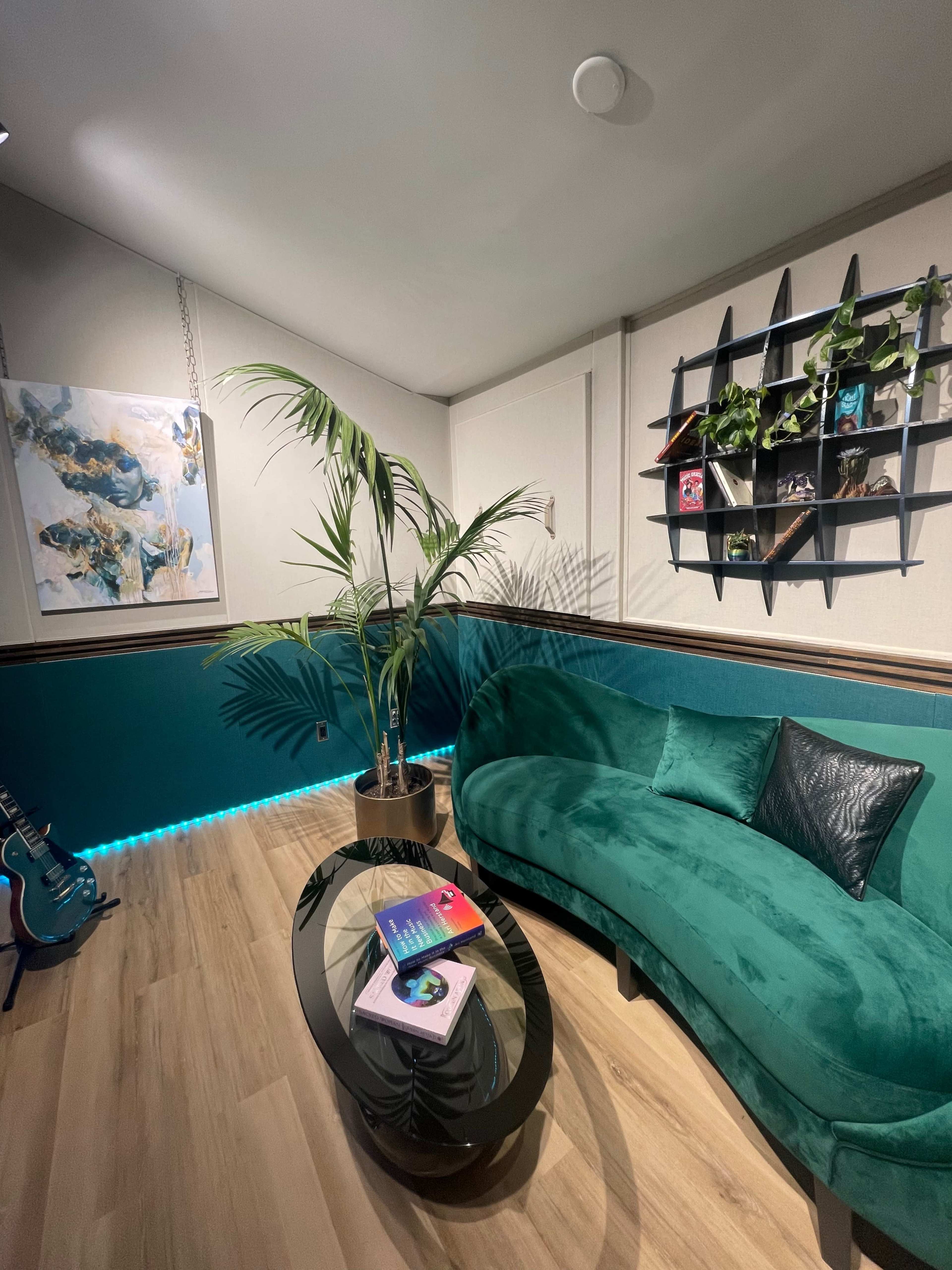A modern lounge area featuring a curved green couch, a glass coffee table with books, a potted palm plant, a wall-mounted shelf with decorative items, and a blue accent wall.
