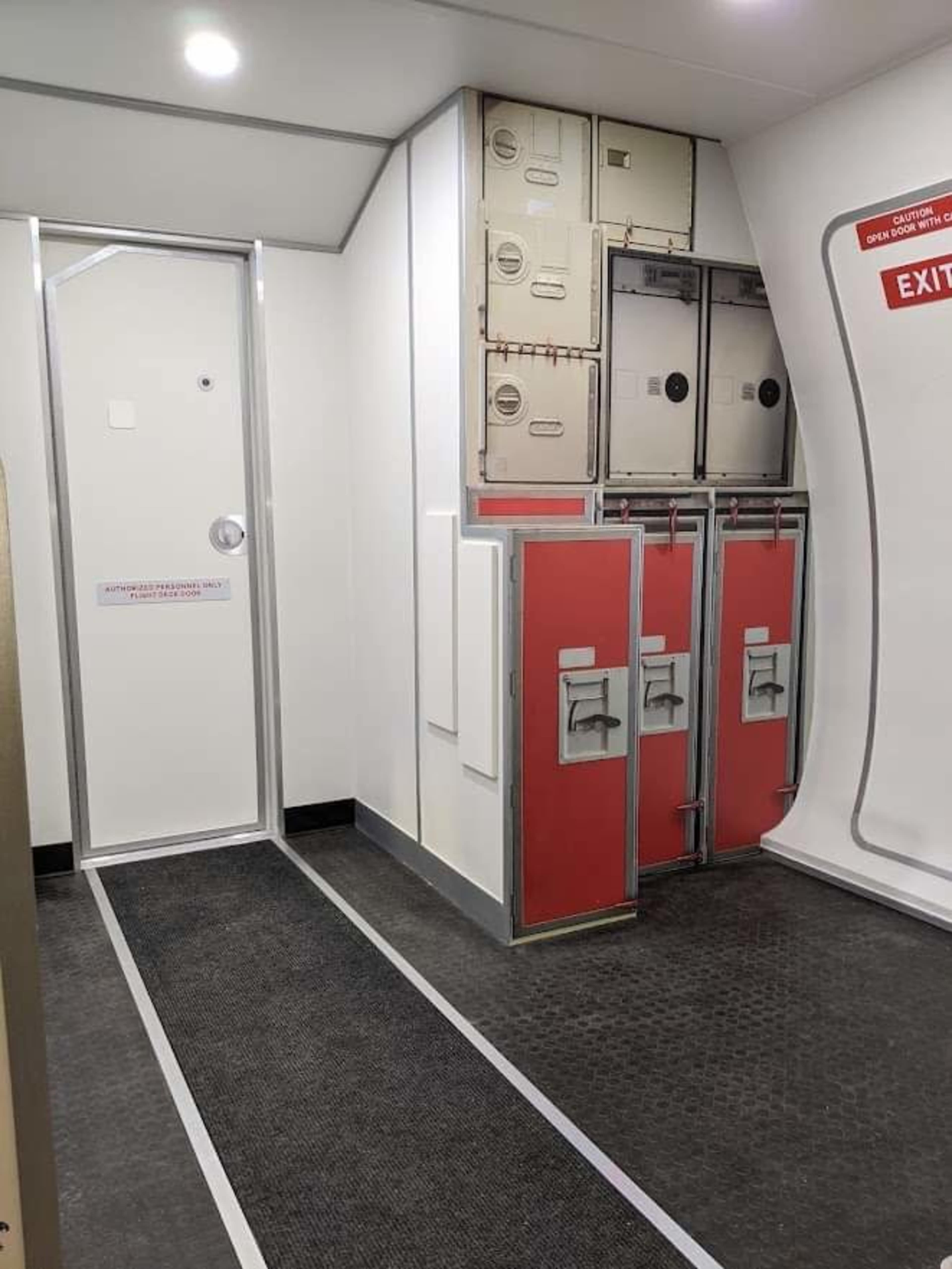 The interior of an airplane, featuring an exit door and several storage compartments with red and gray panels.