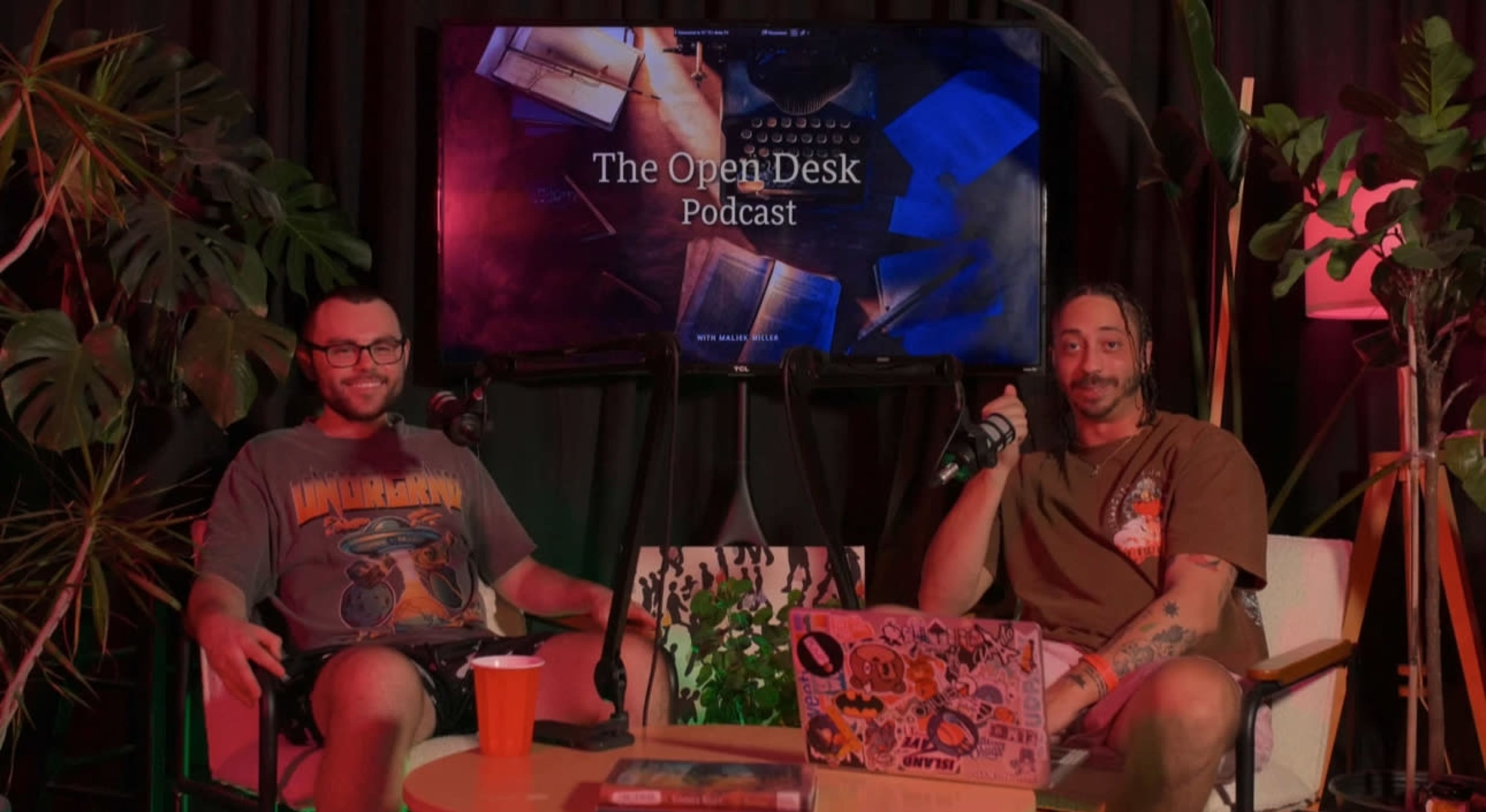 The image features two hosts sitting in a podcast studio with a colorful backdrop and a large screen displaying "The Open Desk Podcast."