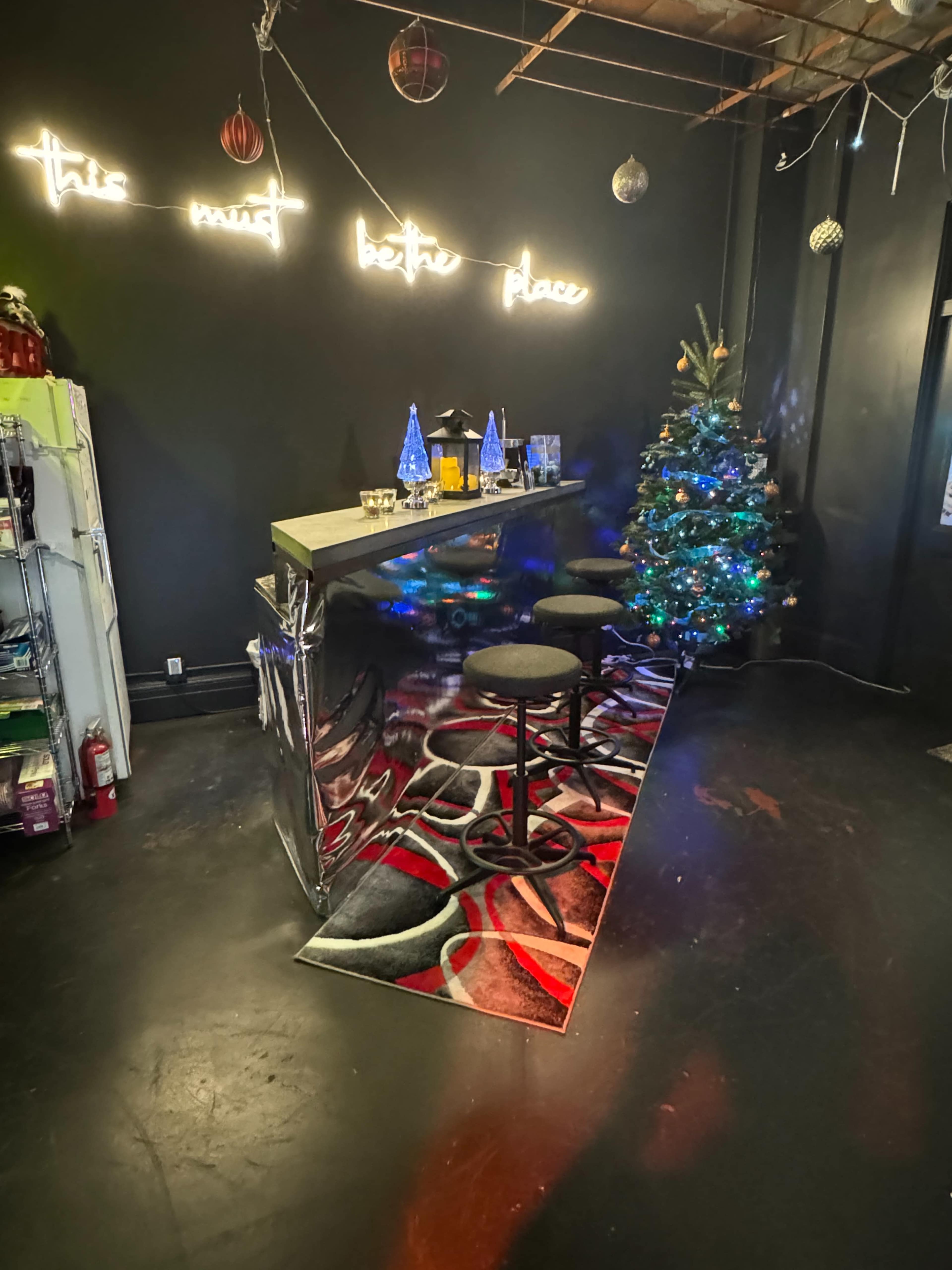 The image shows a small bar area with a metallic counter, three black stools, a decorated Christmas tree, and a neon sign on the wall that reads "this must be a better place."