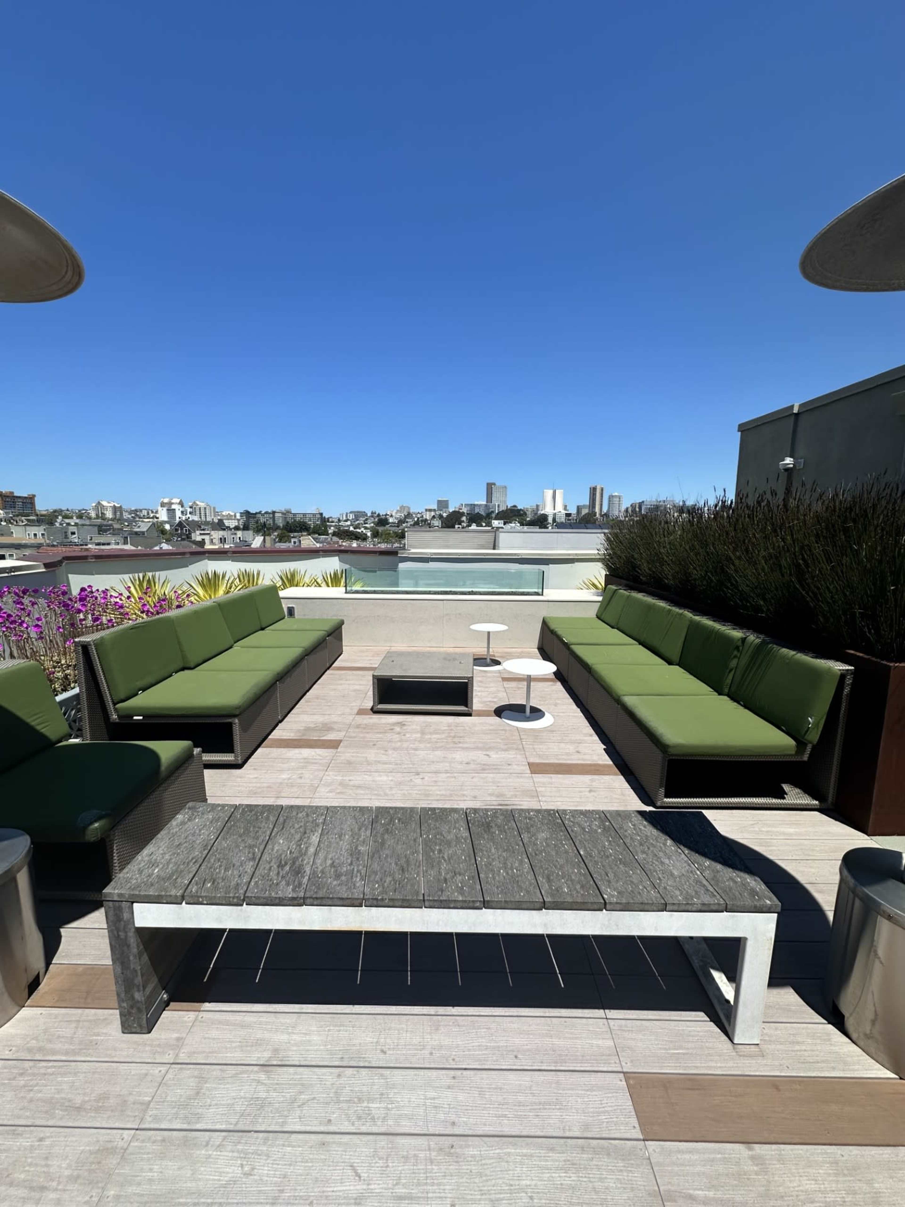 A rooftop terrace features green couches arranged around a central table, with urban buildings visible in the background under a clear blue sky.