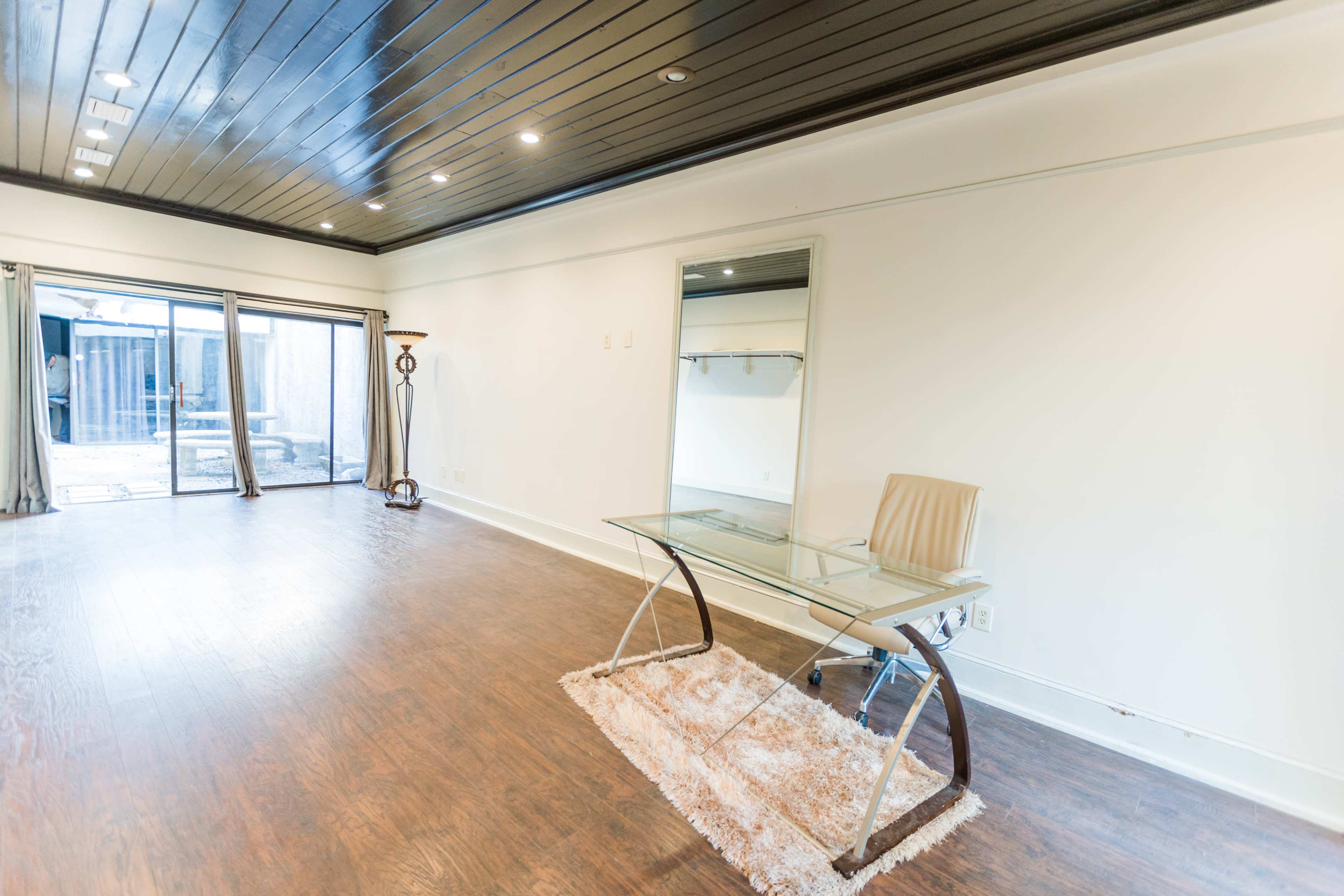 A spacious room with a glass-top desk and a chair on a rug, featuring large windows that lead to an outdoor area.