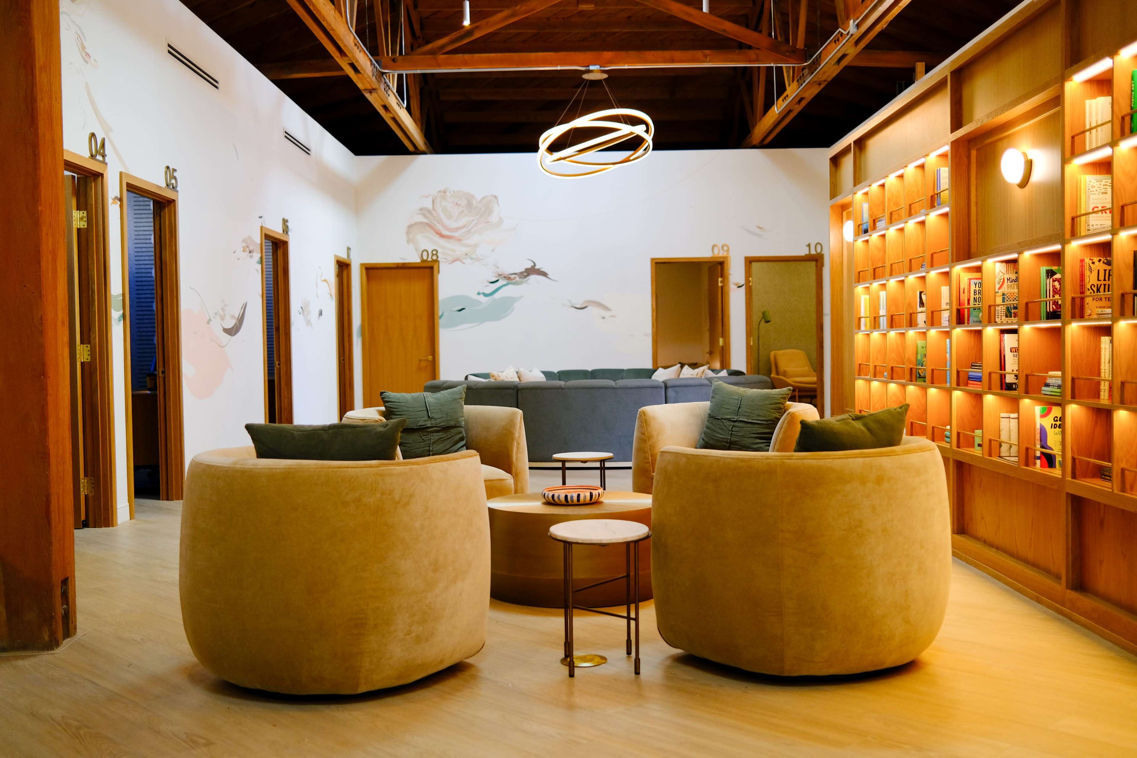 The image shows a cozy interior with two round, plush chairs facing a wooden table, surrounded by warm-toned walls and shelves filled with books.
