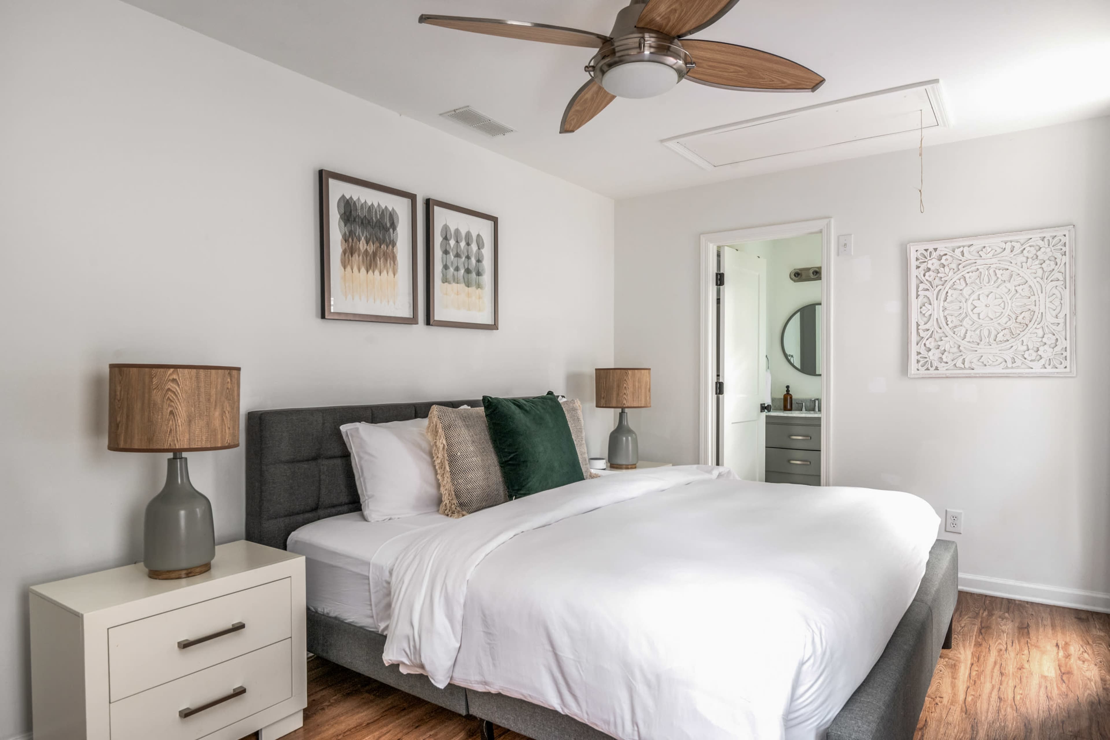 The image shows a well-lit bedroom featuring a gray bed with a white comforter, two bedside tables with lamps, framed artwork on the wall, and a ceiling fan.