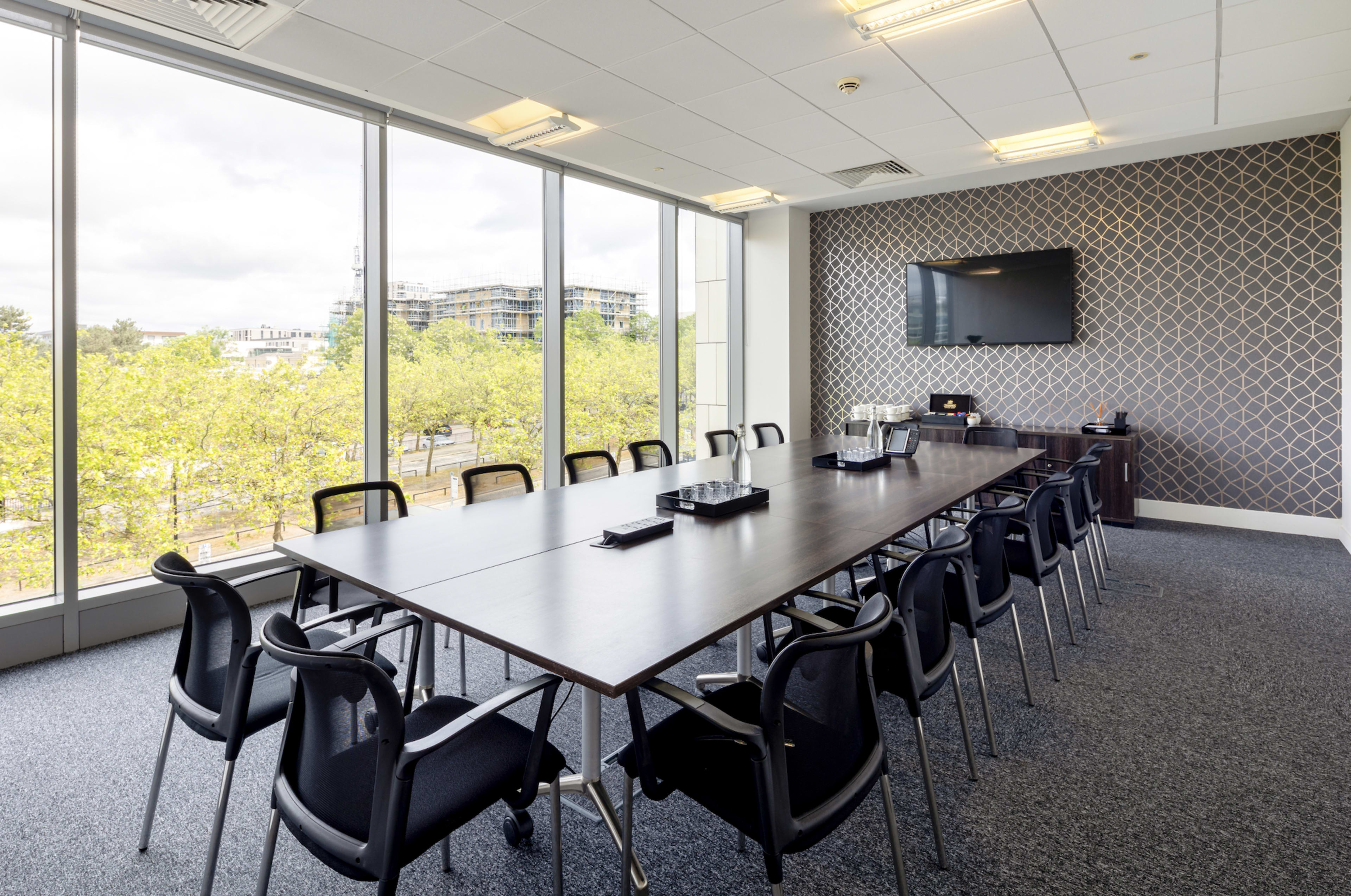 The image features a conference room with a large rectangular table surrounded by black chairs, large windows overlooking greenery, and a wall-mounted television.