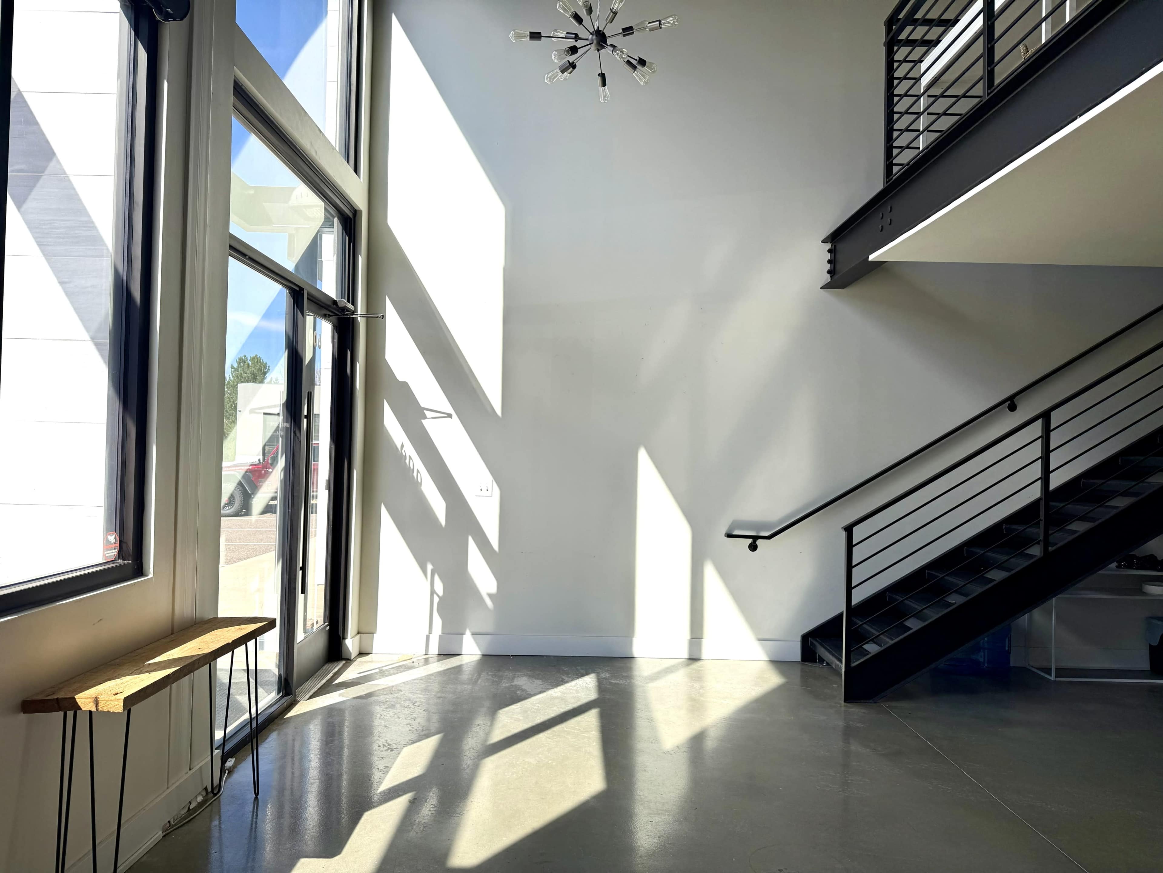 The image shows a spacious interior with a large window, a wooden bench against the wall, and a staircase leading to an upper level.