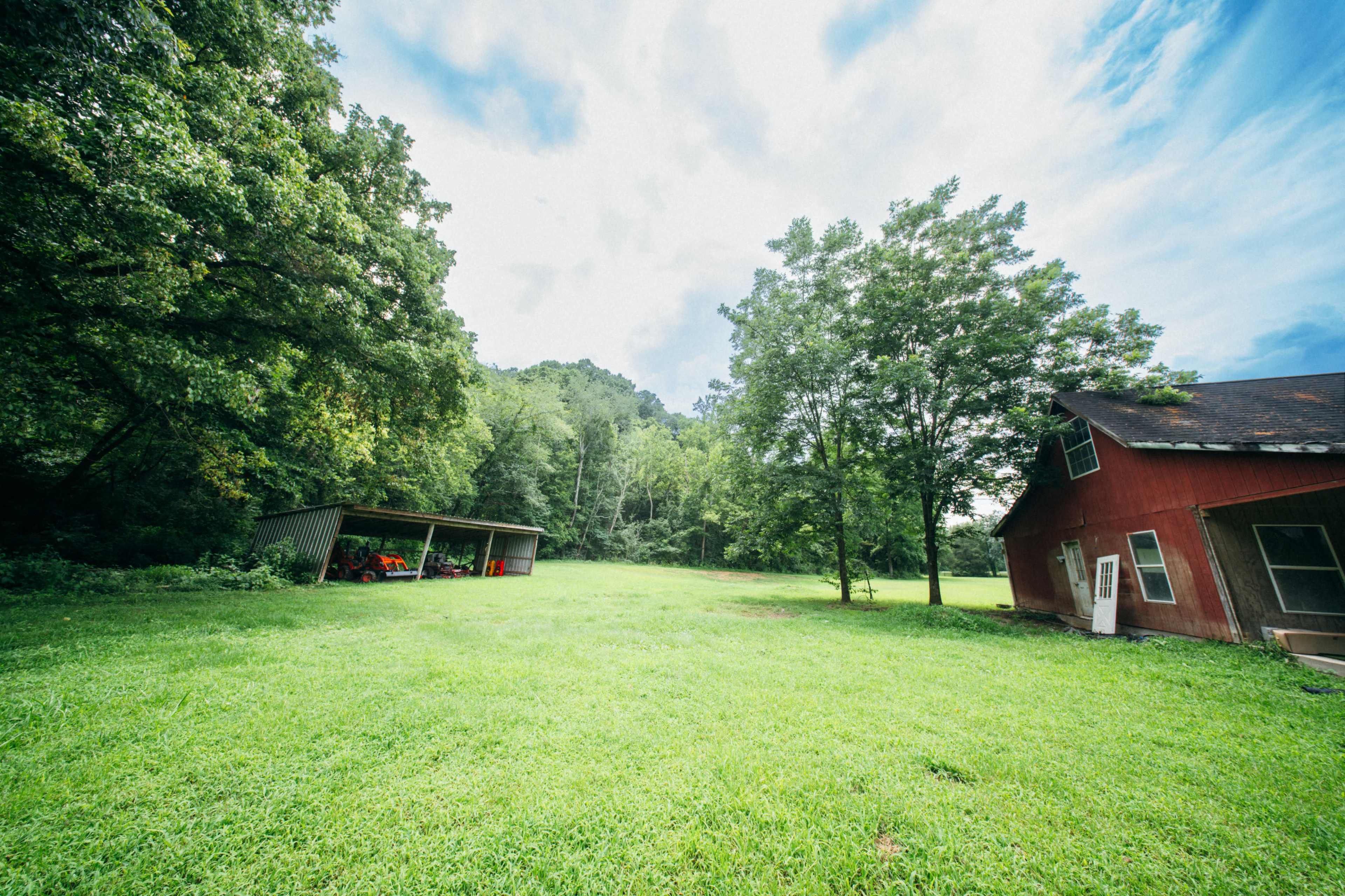 A red barn is situated near a lush green field surrounded by trees under a partly cloudy sky.