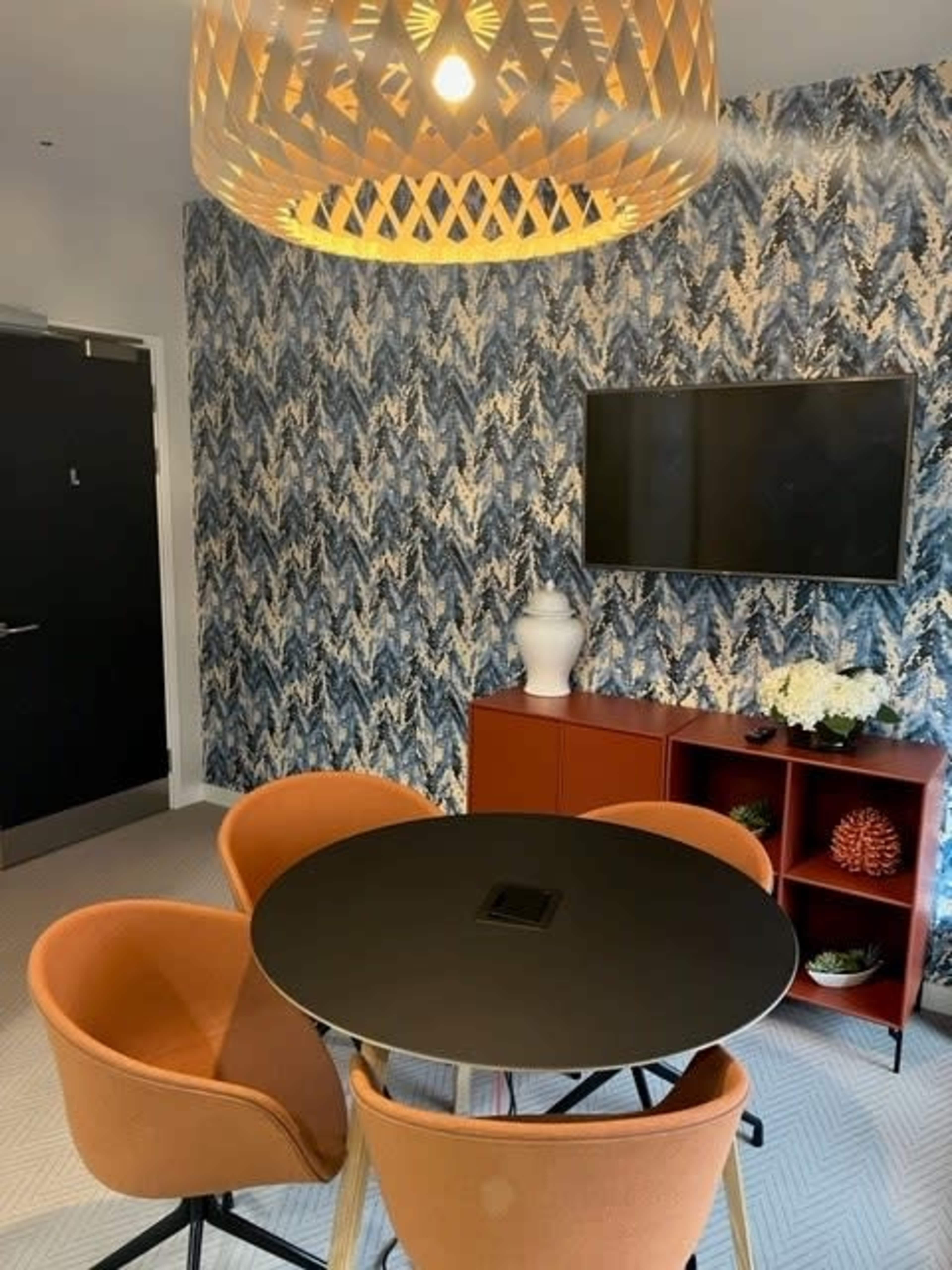 A modern meeting room features a round black table surrounded by orange chairs, a patterned blue wallpaper, and a television mounted on the wall.