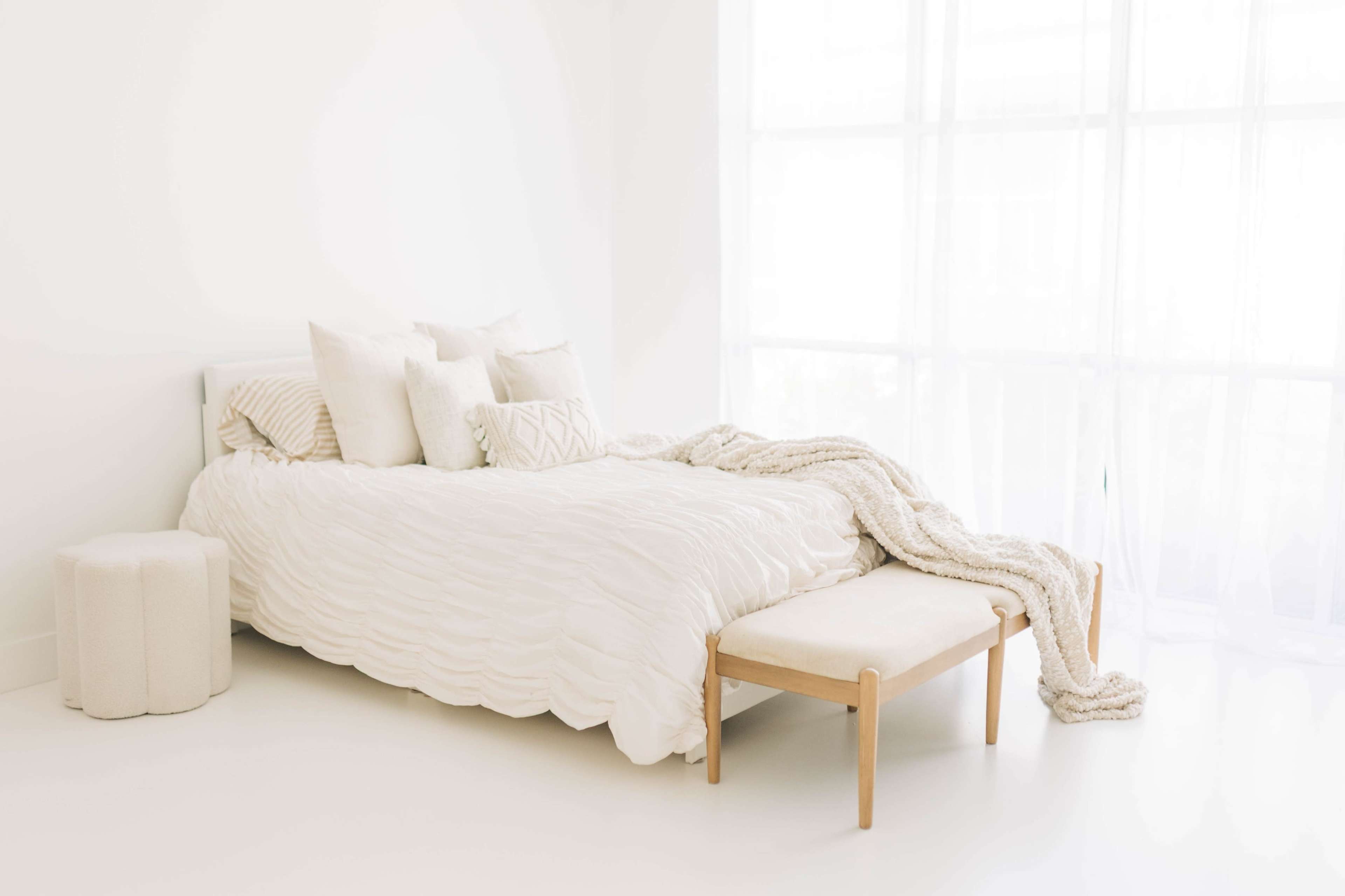 The image shows a minimalist bedroom featuring a white bed with several pillows, a light-colored bench at its foot, and a textured blanket draped over the side.