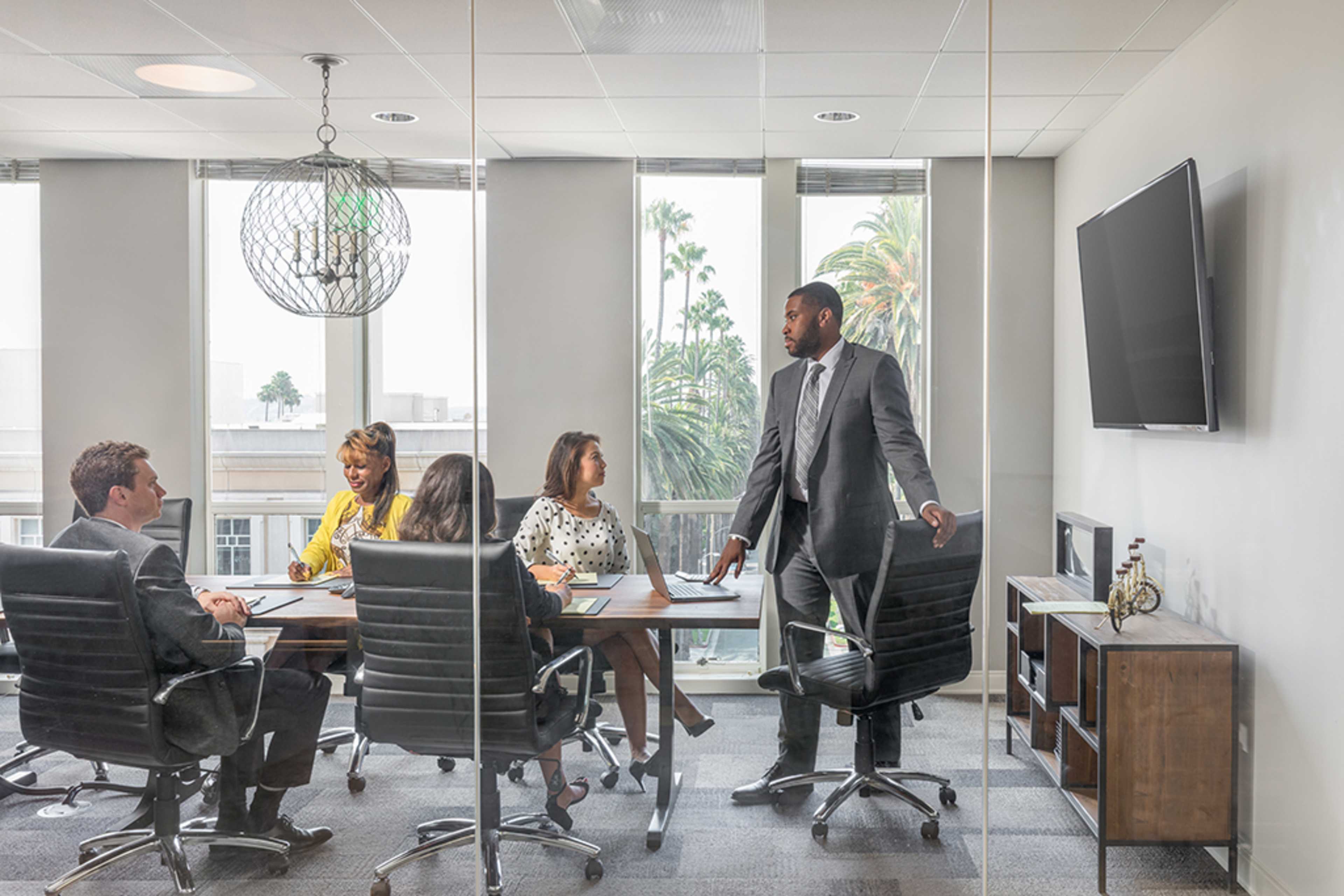 A group of six professionals is seated around a conference table in a modern office with large windows overlooking palm trees.
