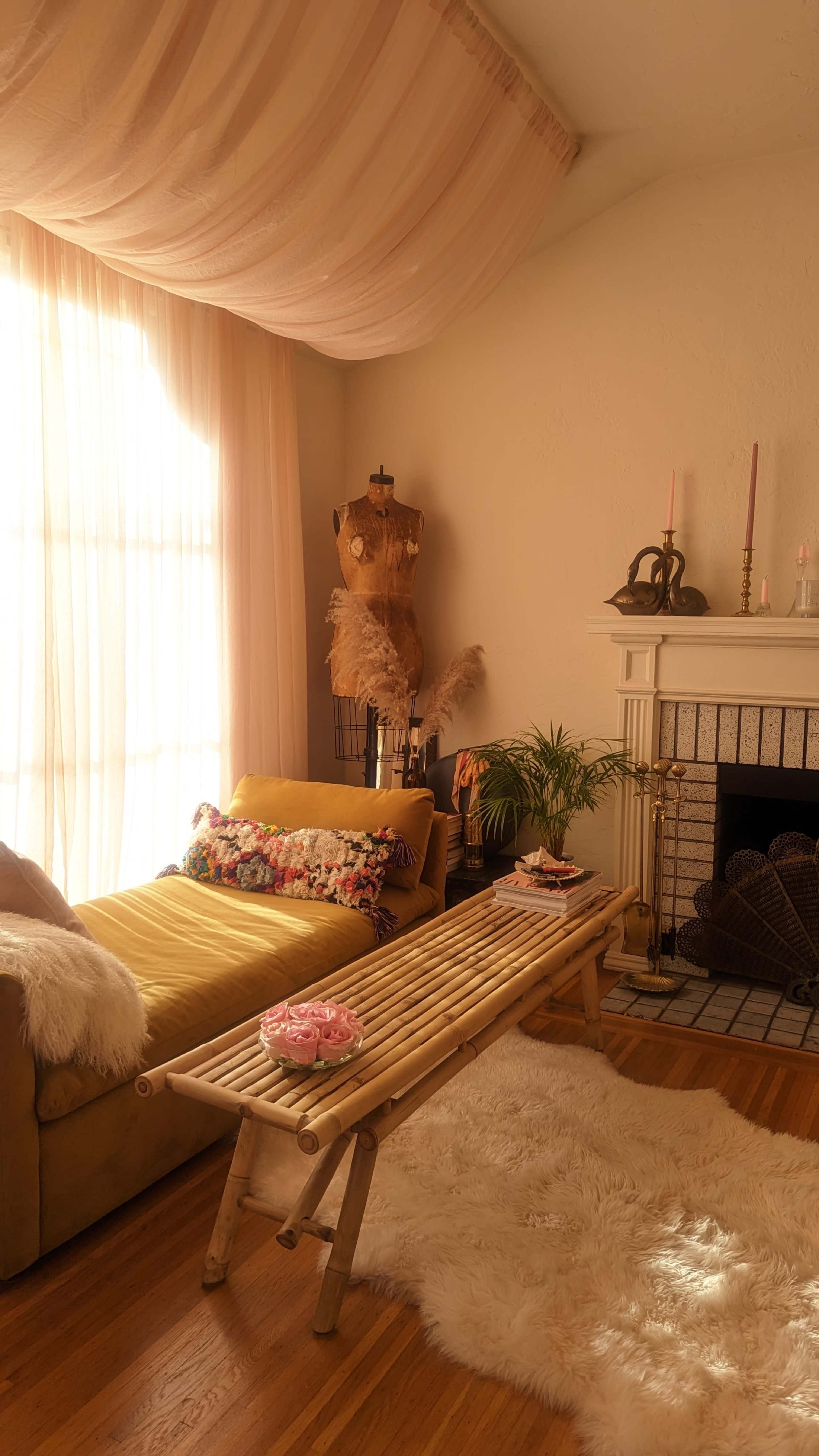 The scene shows a cozy room with a yellow daybed, a wooden table, a mannequin dressed in fabric, and a fireplace flanked by candles.