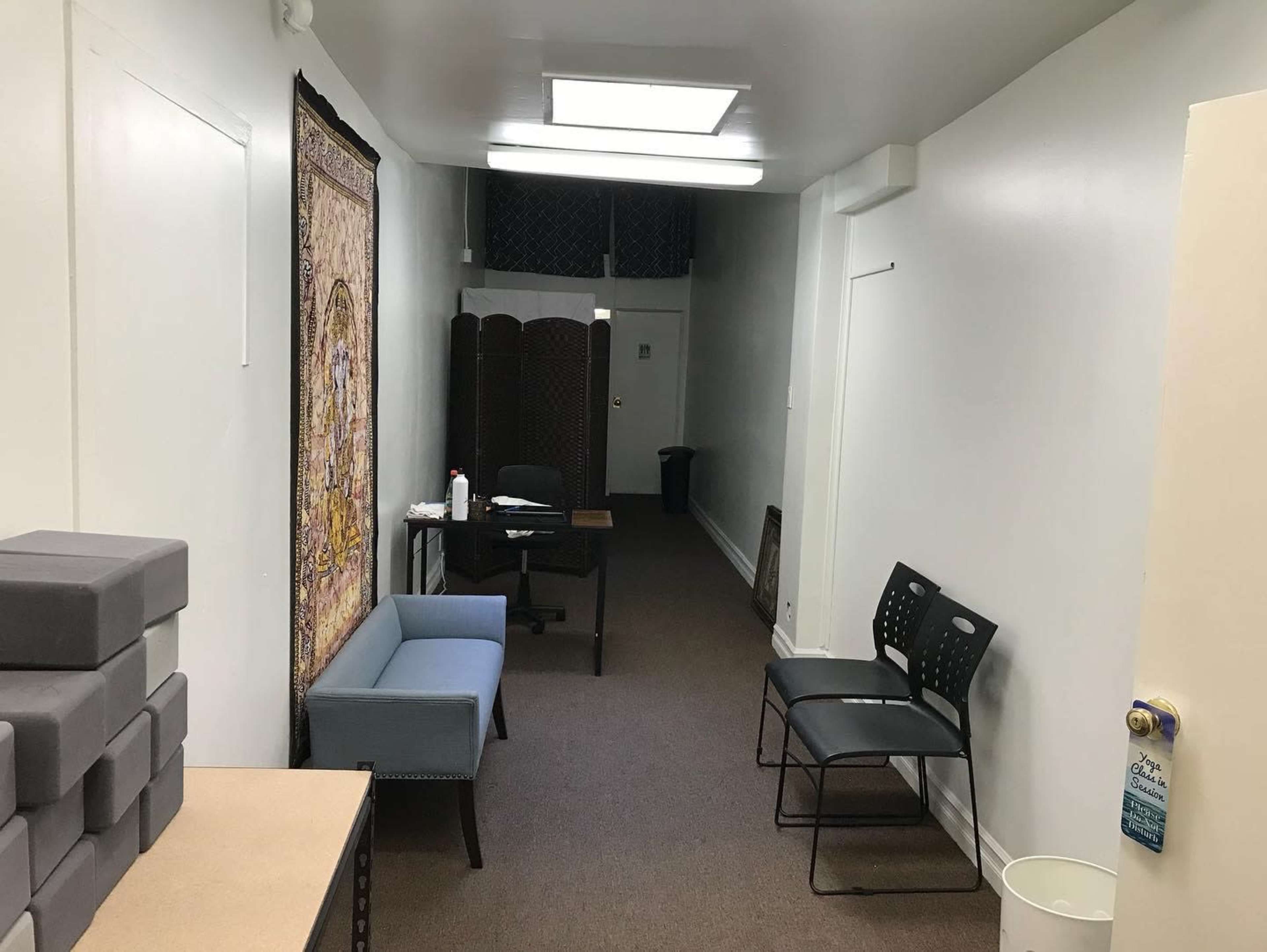 The image shows a narrow hallway with a carpeted floor, featuring a small table with a chair, two chairs against the wall, a decorative tapestry, and a wastebasket at the end.