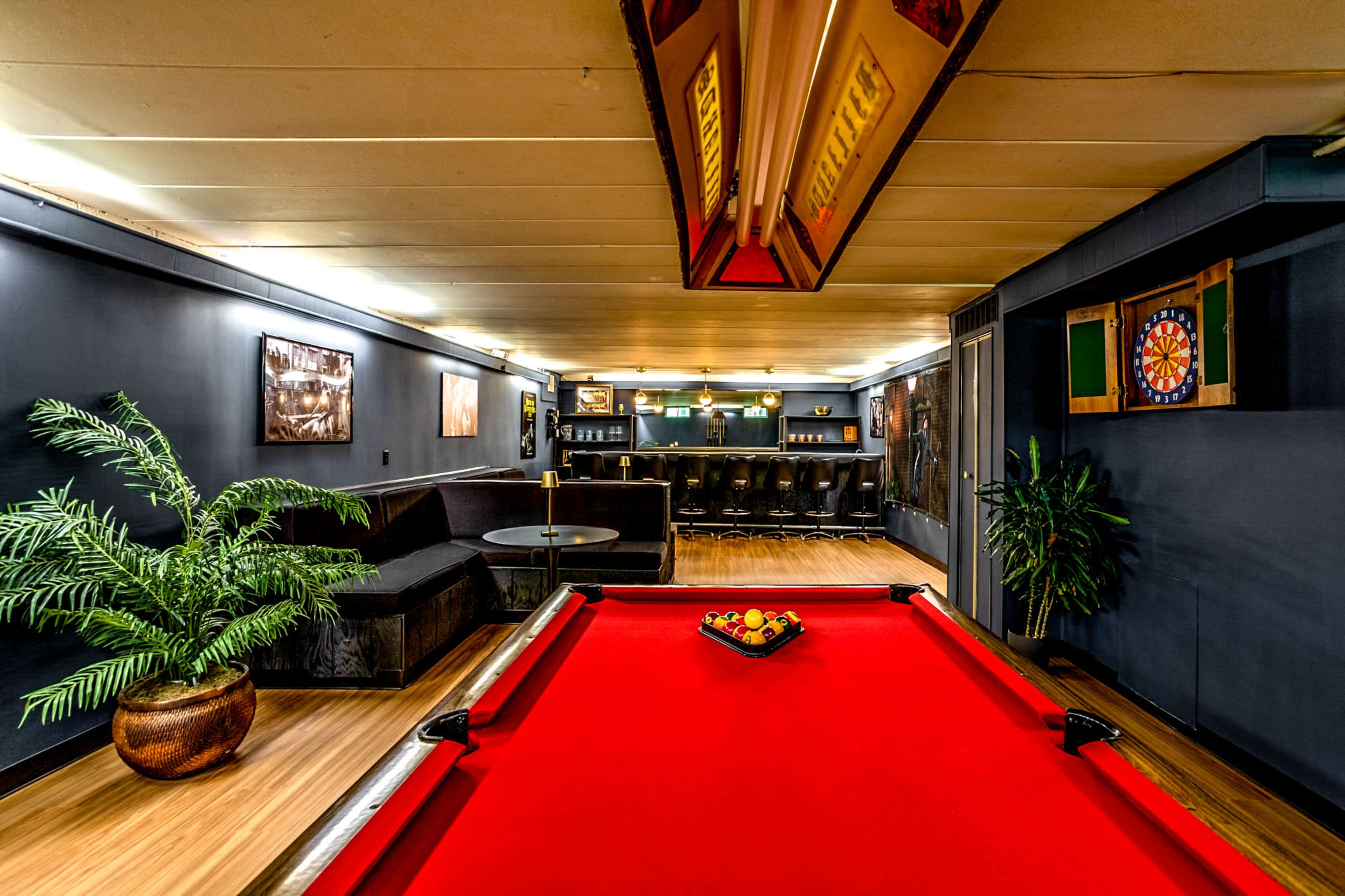 A game room featuring a red pool table, a bar area with stools, and a lounge space with a large sofa and decorative plants.