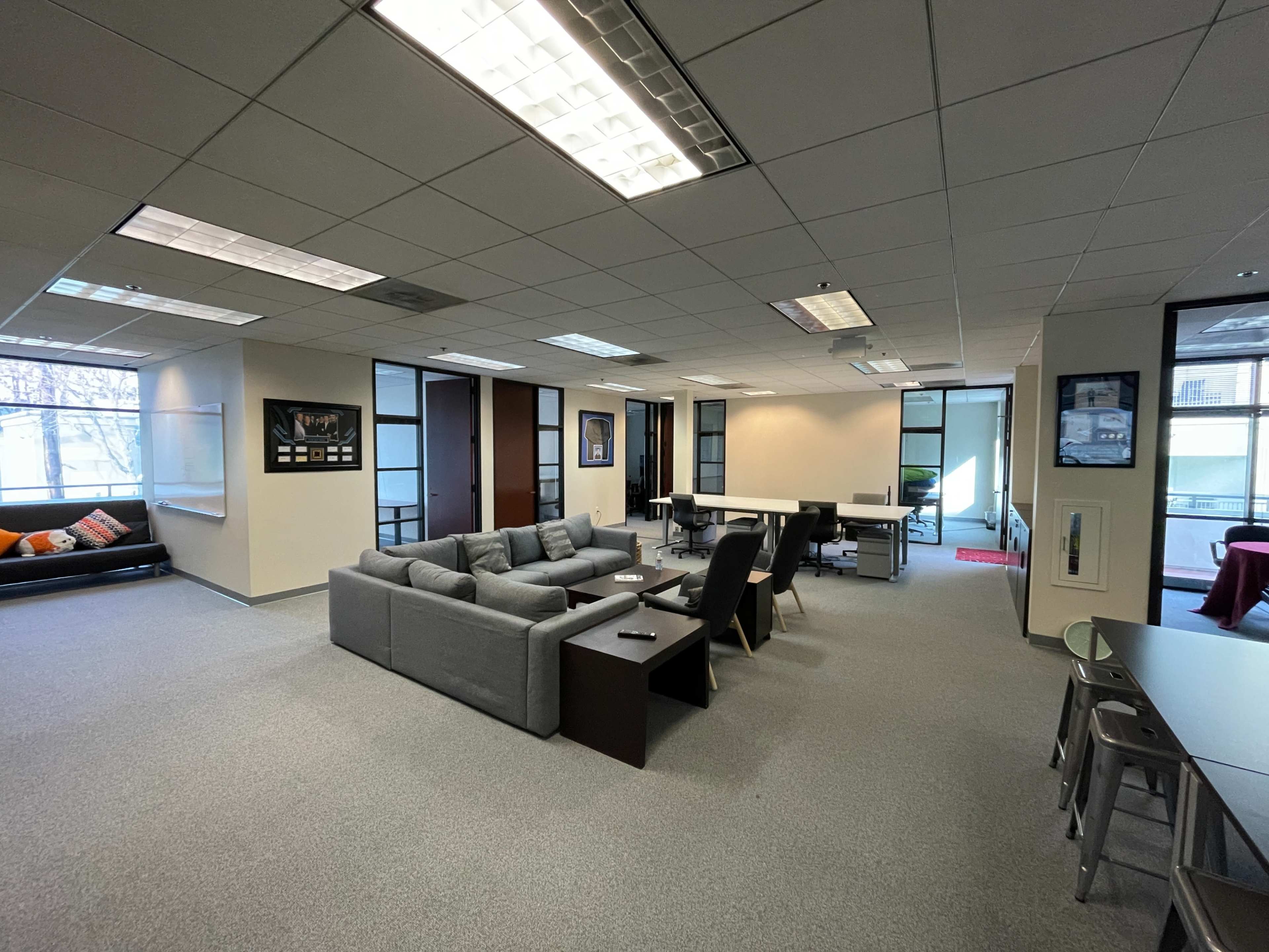 The image shows a modern office space featuring a lounge area with a gray sofa and a coffee table, along with desks and chairs in the surrounding workstations.