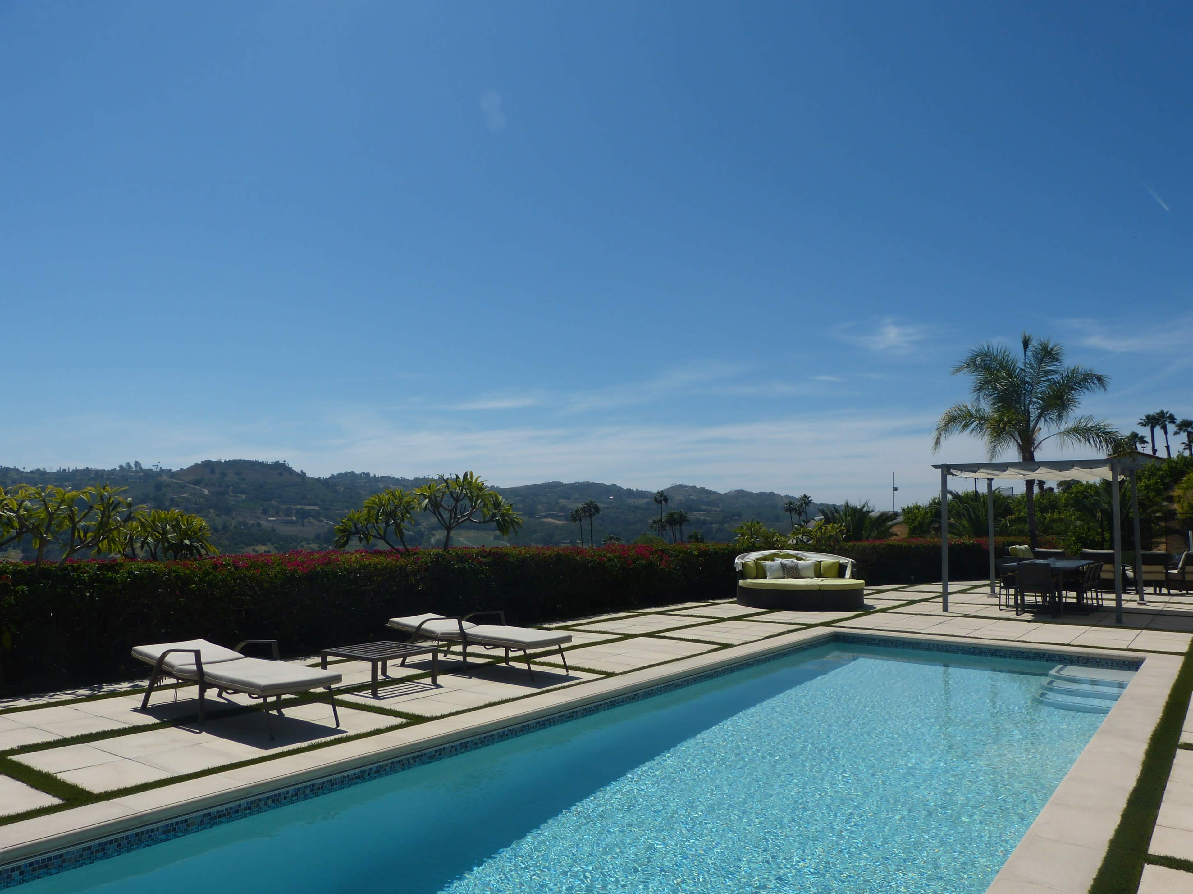 A clear blue sky overlooks a swimming pool surrounded by lounge chairs and landscaped greenery with hills in the background.