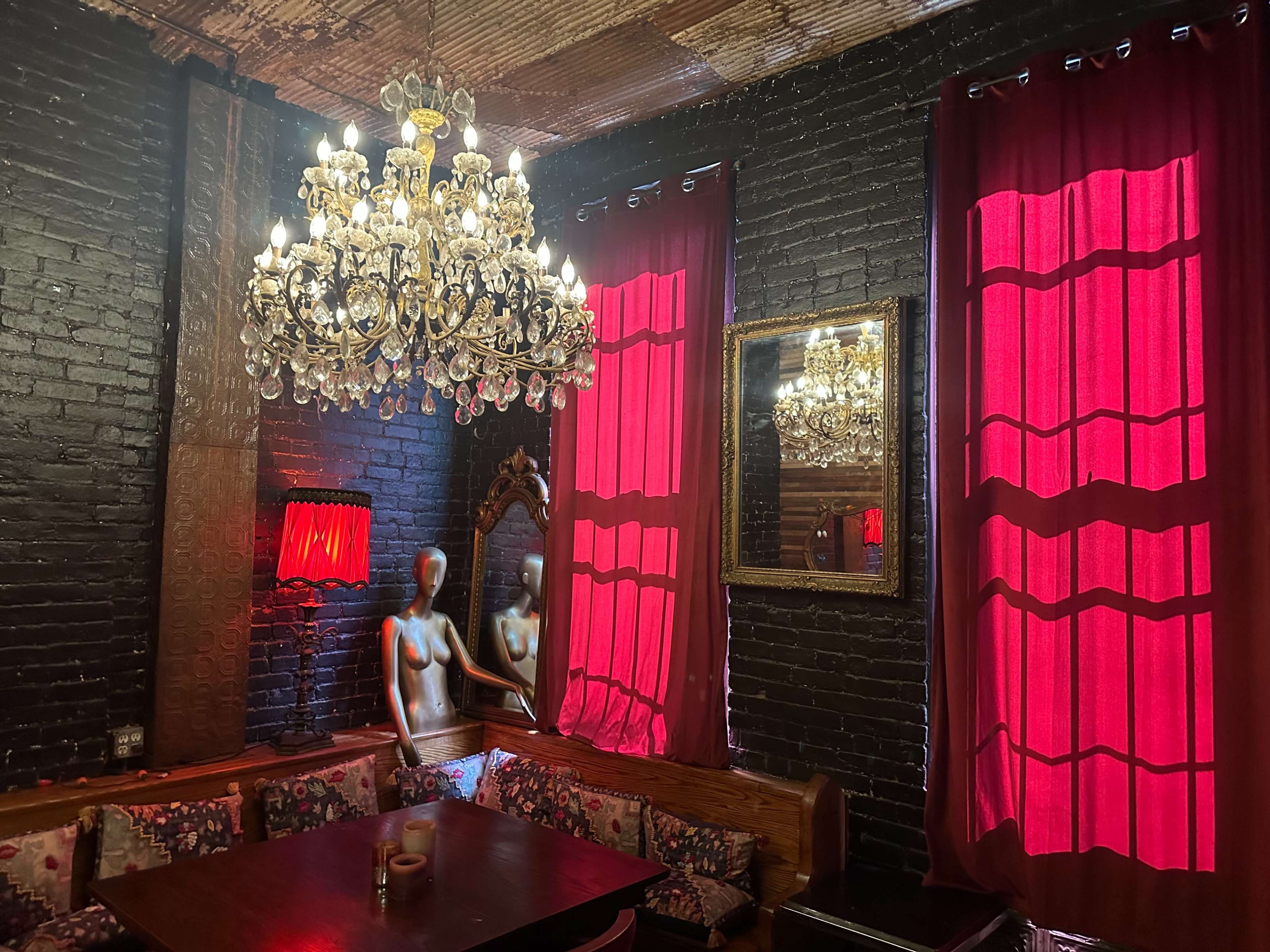 The image shows a dimly lit room with black brick walls, red curtains, a chandelier, and a mirrored wall featuring a mannequin.