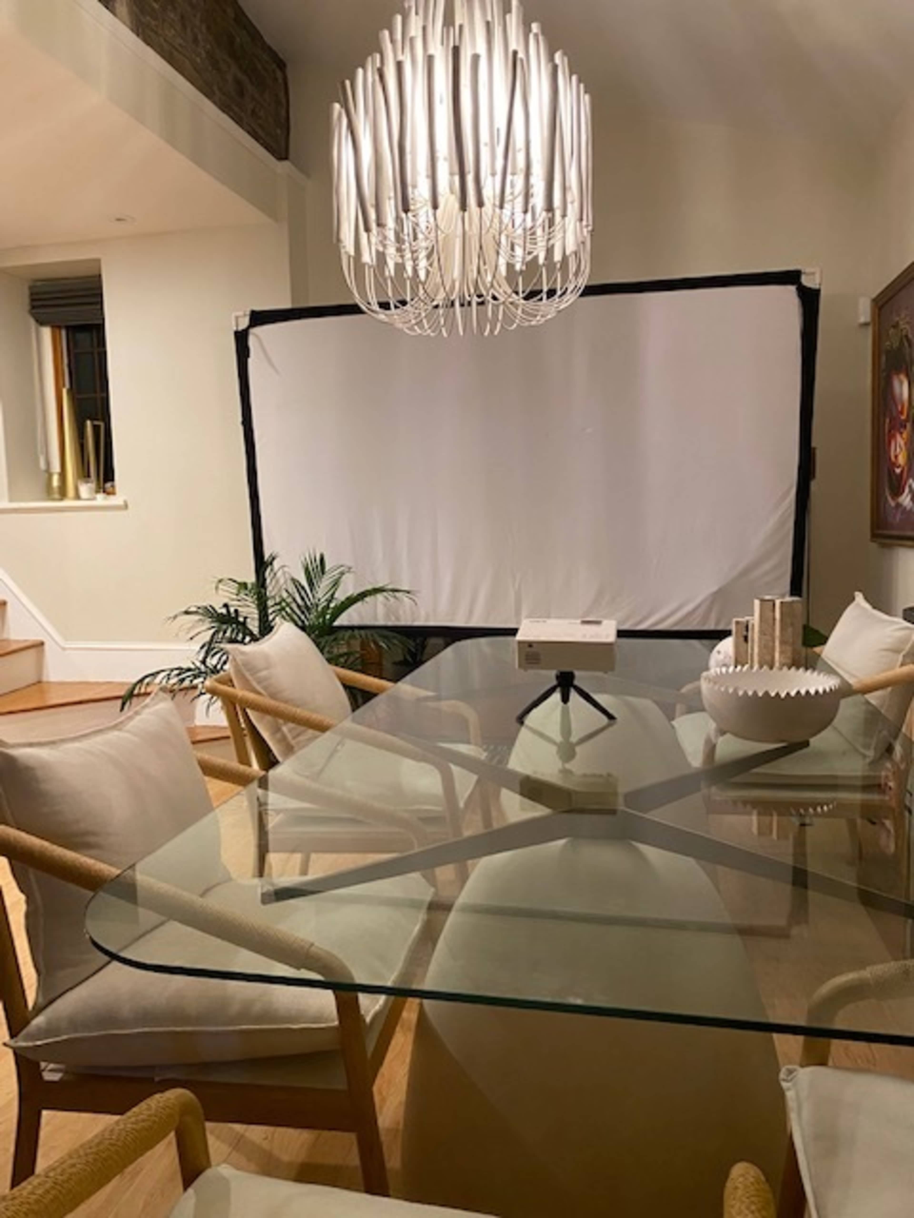 A dining room features a glass table surrounded by four chairs, with a large chandelier overhead and a blank projection screen in the background.
