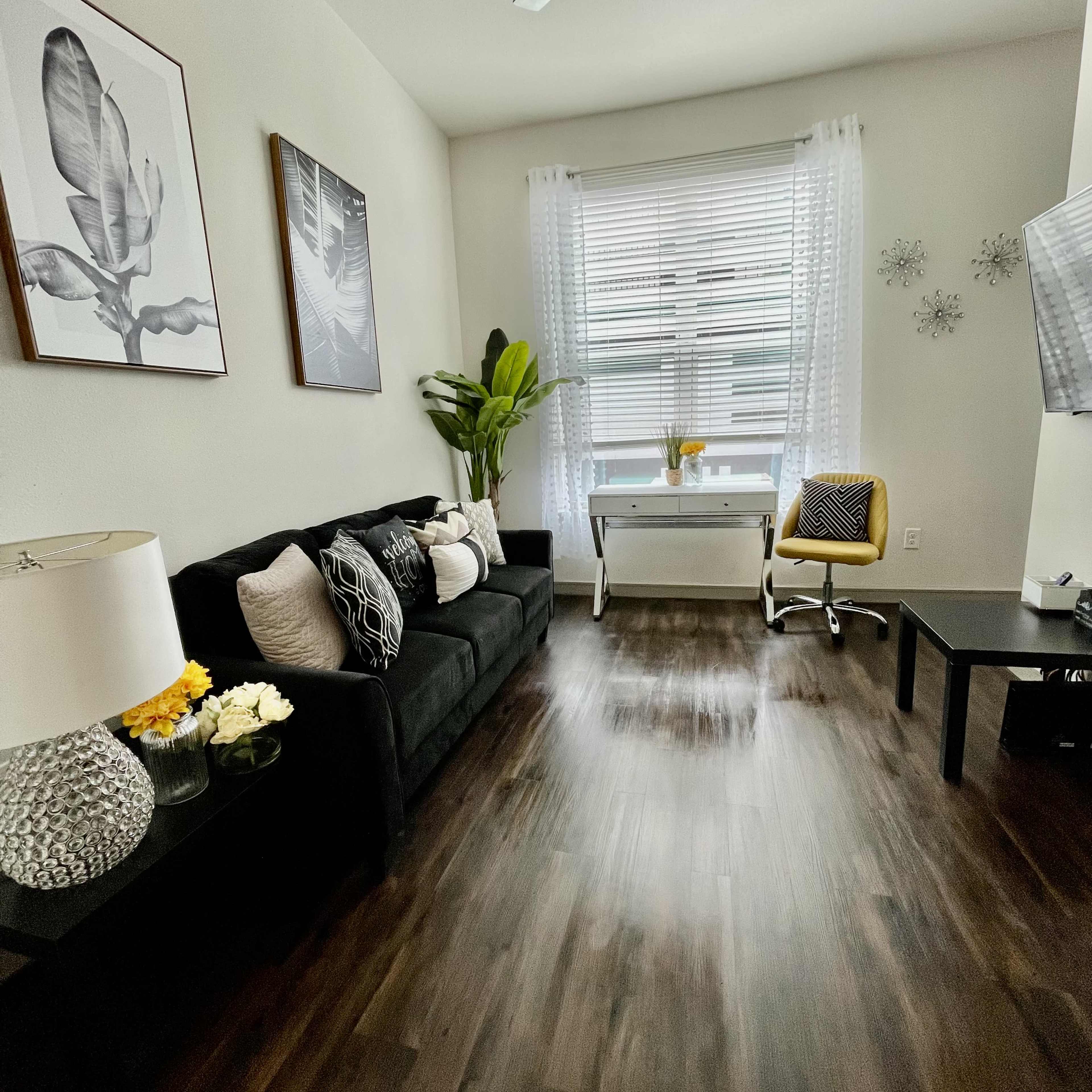 The room features a black sofa, a small desk with a chair, and artwork on the walls, complemented by a large window with sheer curtains.