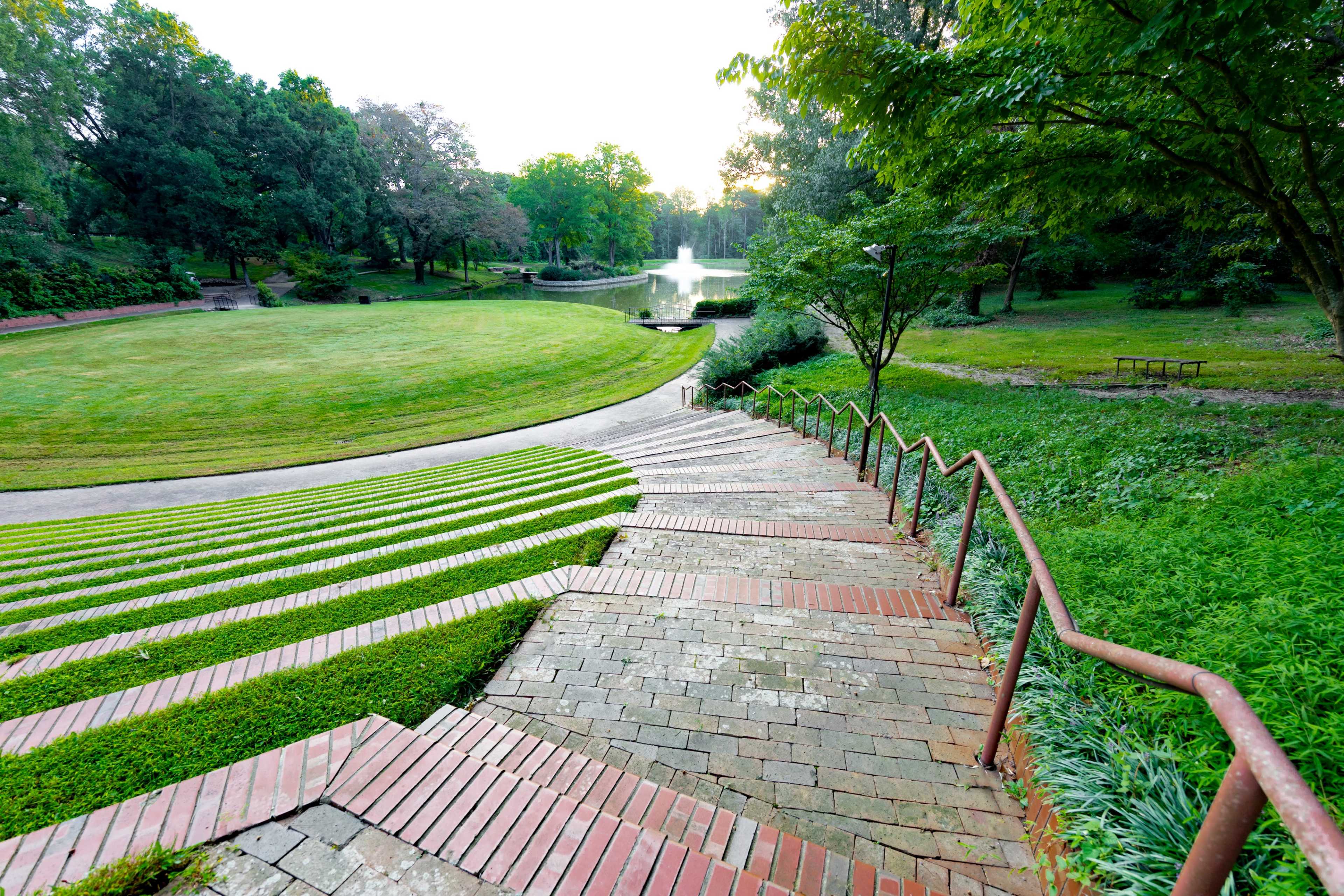 A curved brick walkway descends a grassy slope towards a pond surrounded by trees.