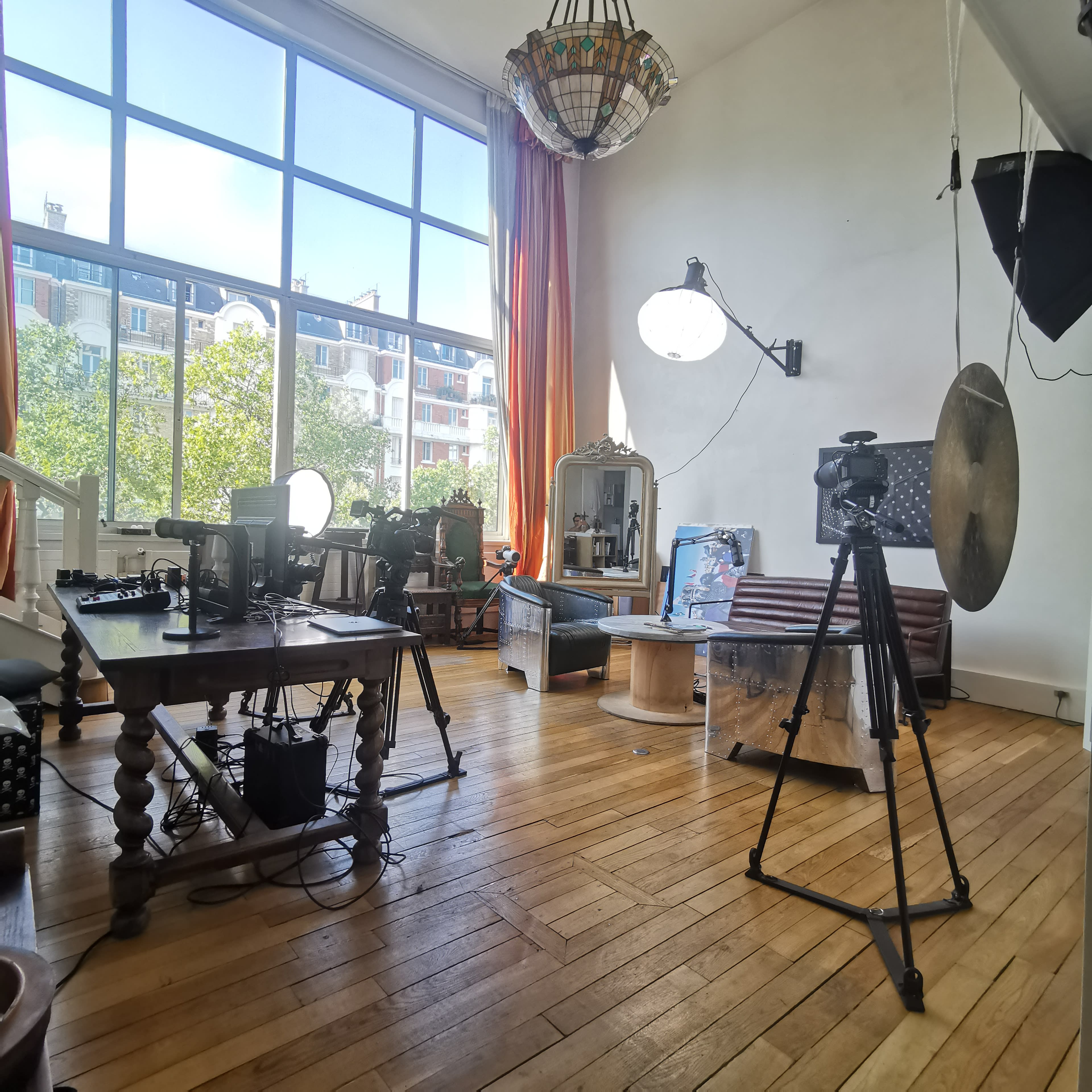 A bright room with large windows, featuring a wooden table with cameras, lights, and a large cymbal on a stand.