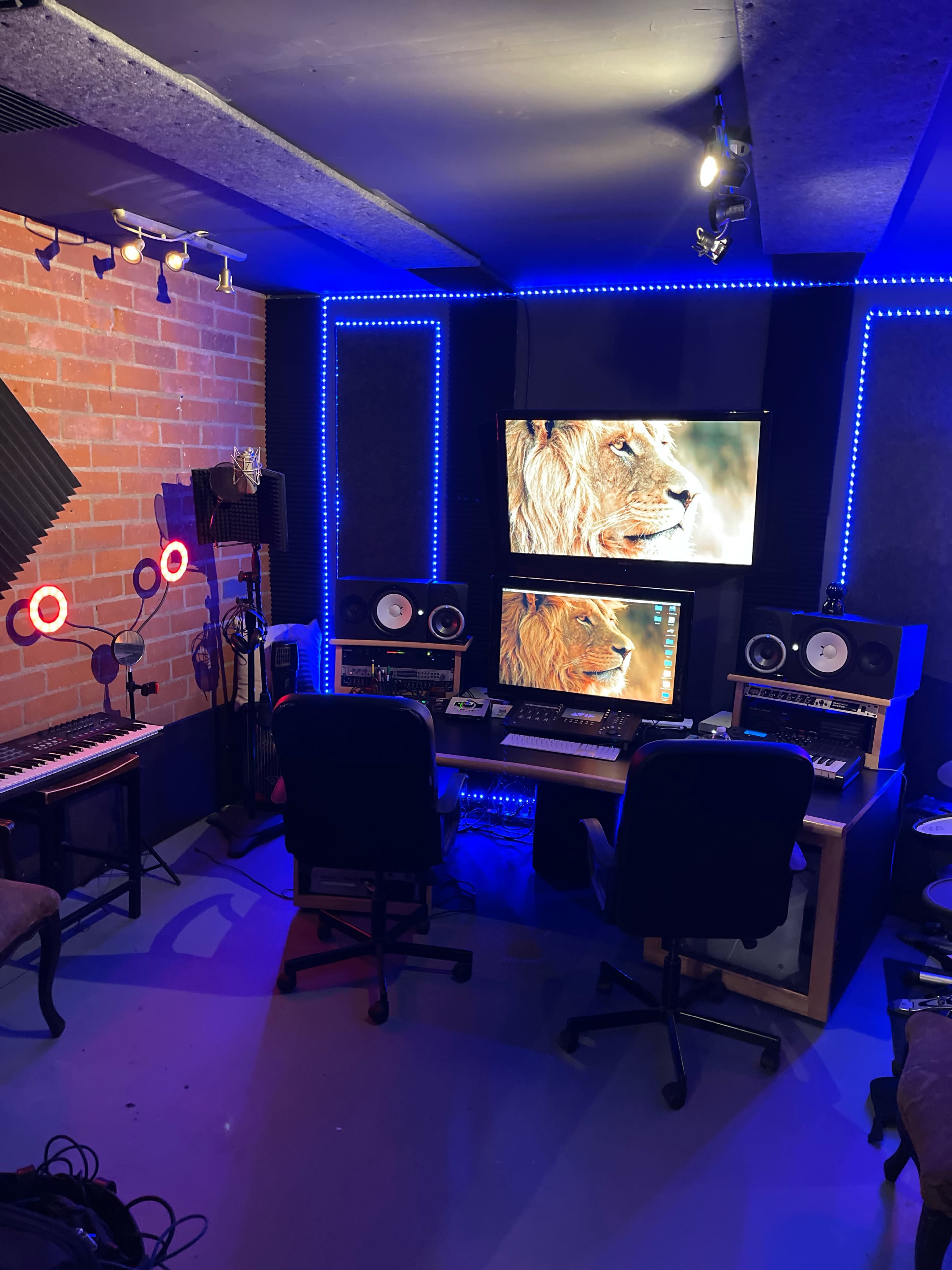 A modern recording studio featuring a desk with two computer monitors, studio monitors, a keyboard, microphones, and colorful LED lighting along the walls.