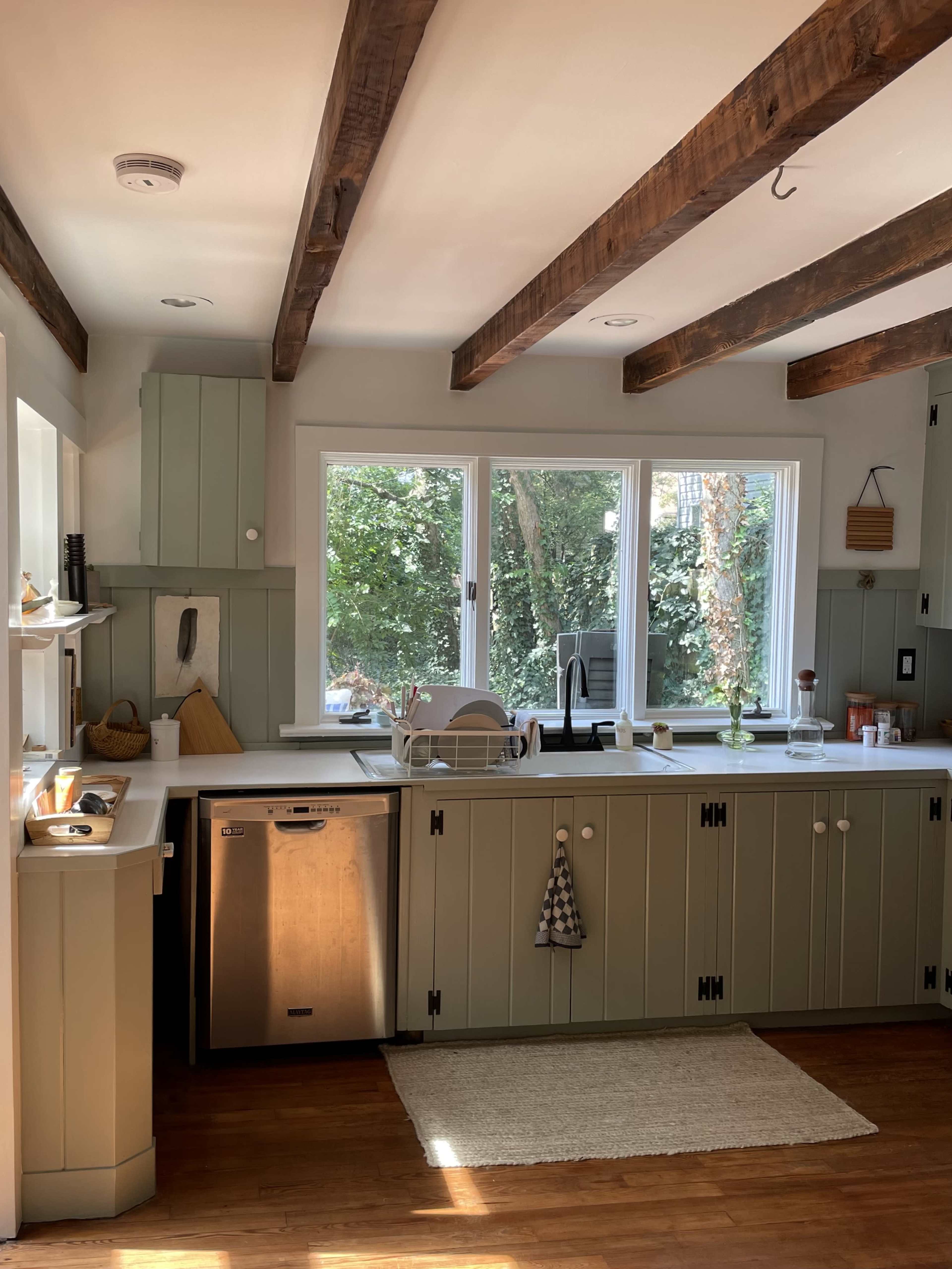 A kitchen features green cabinetry, wooden beams, and a window overlooking a garden.