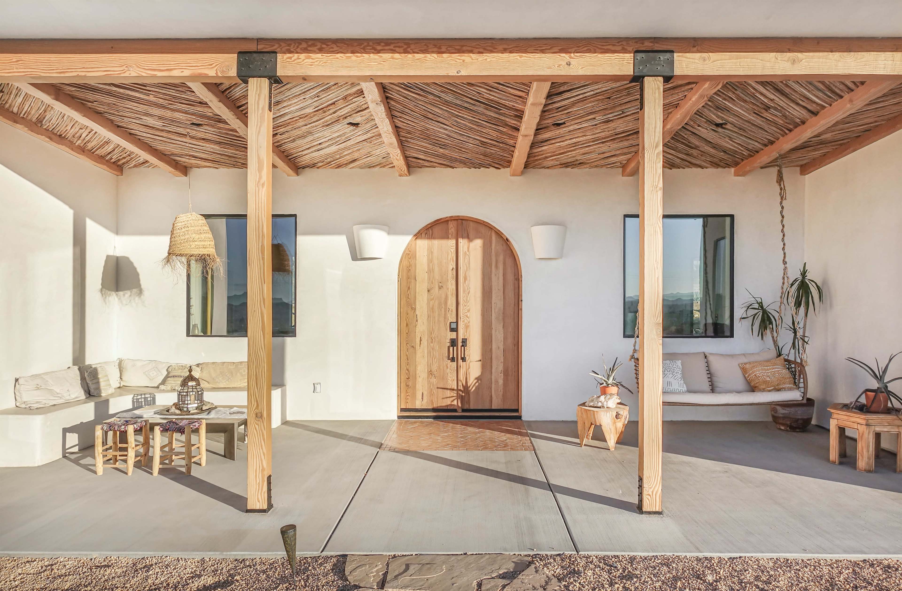 A modern entrance with a wooden door framed by large, open beams, a thatched roof, and minimalist seating arrangements on a concrete patio.