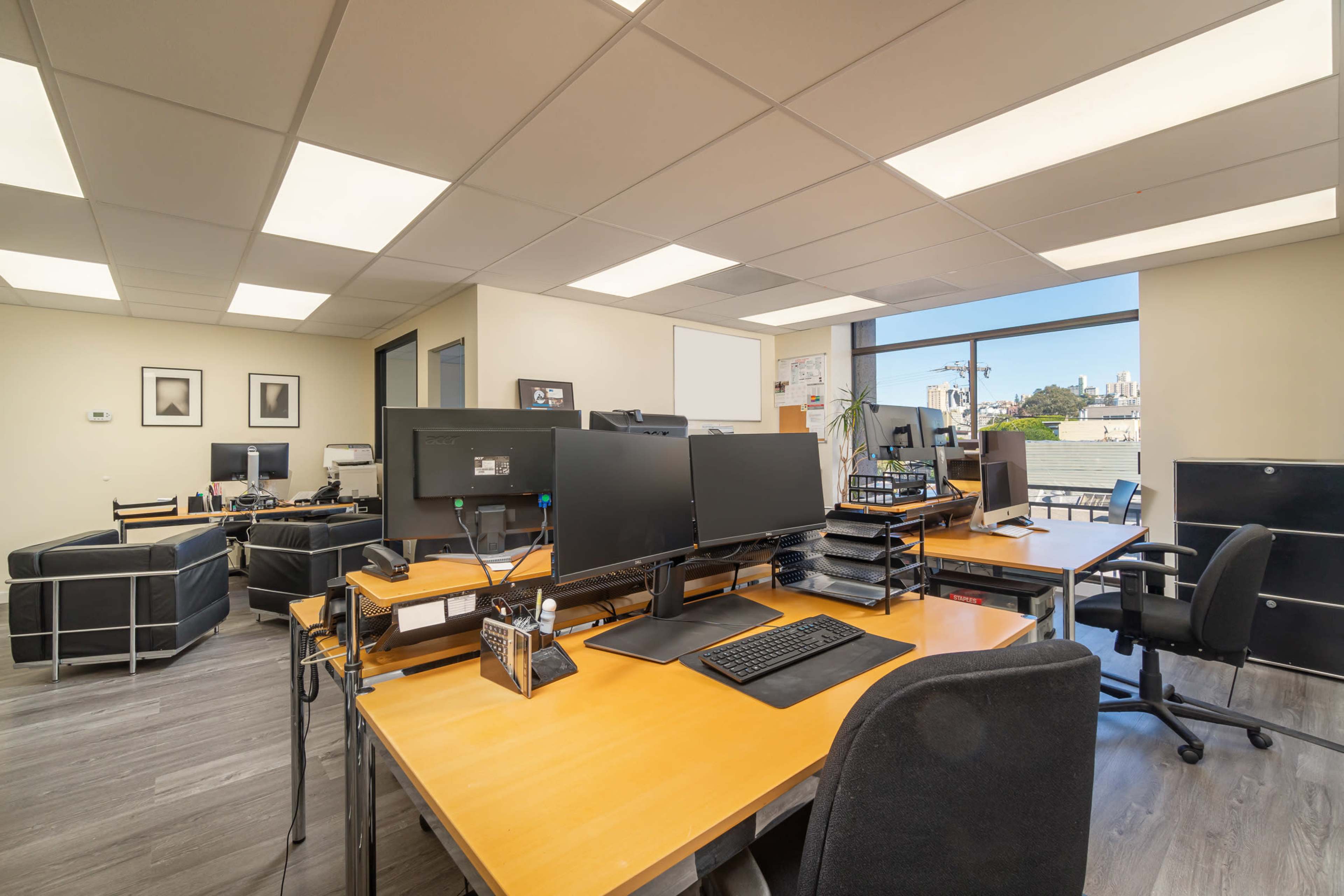 A modern office space features multiple desks with computers, large monitors, and a view of the city through a window.