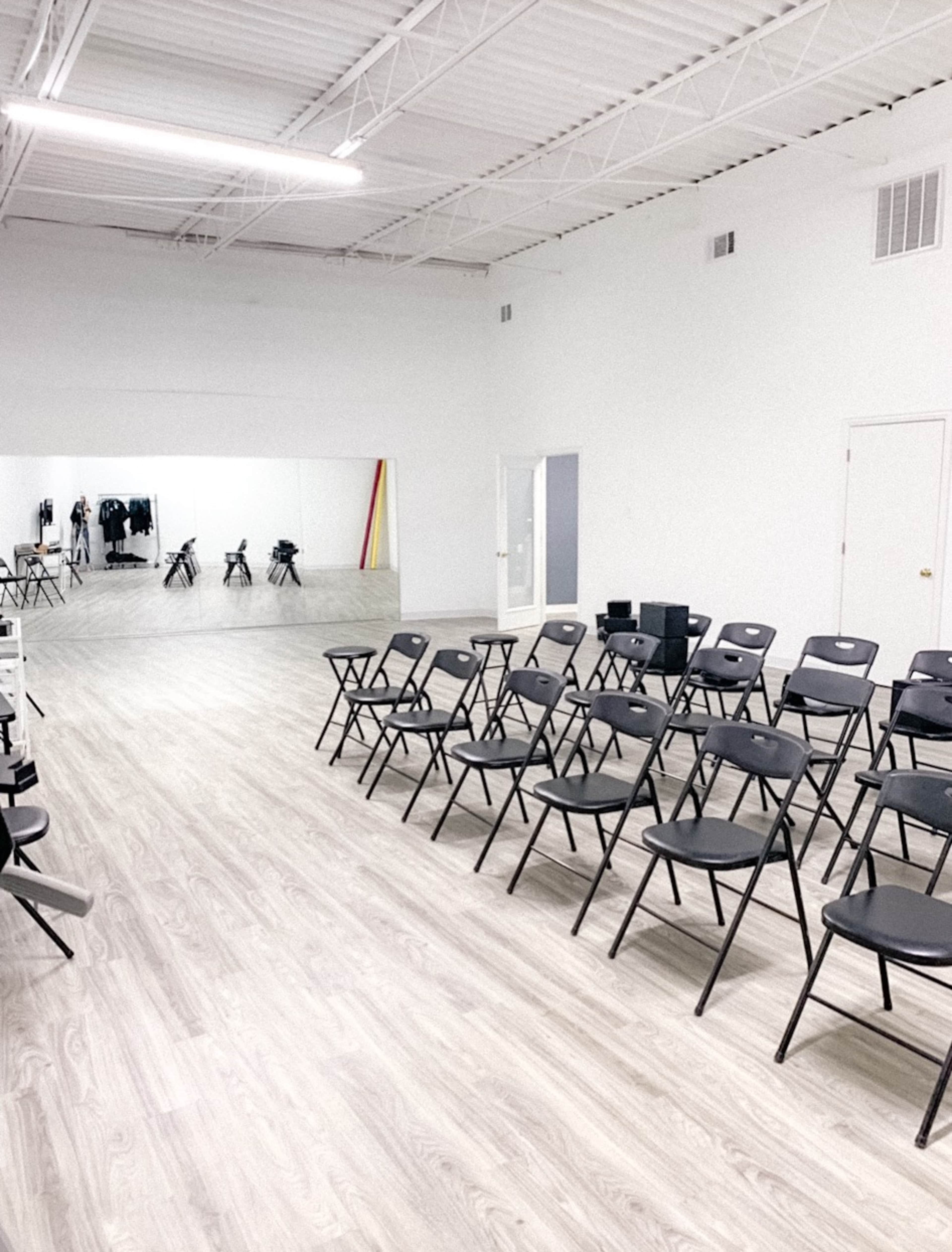 A spacious room with rows of black folding chairs facing a large mirror on a white wall.