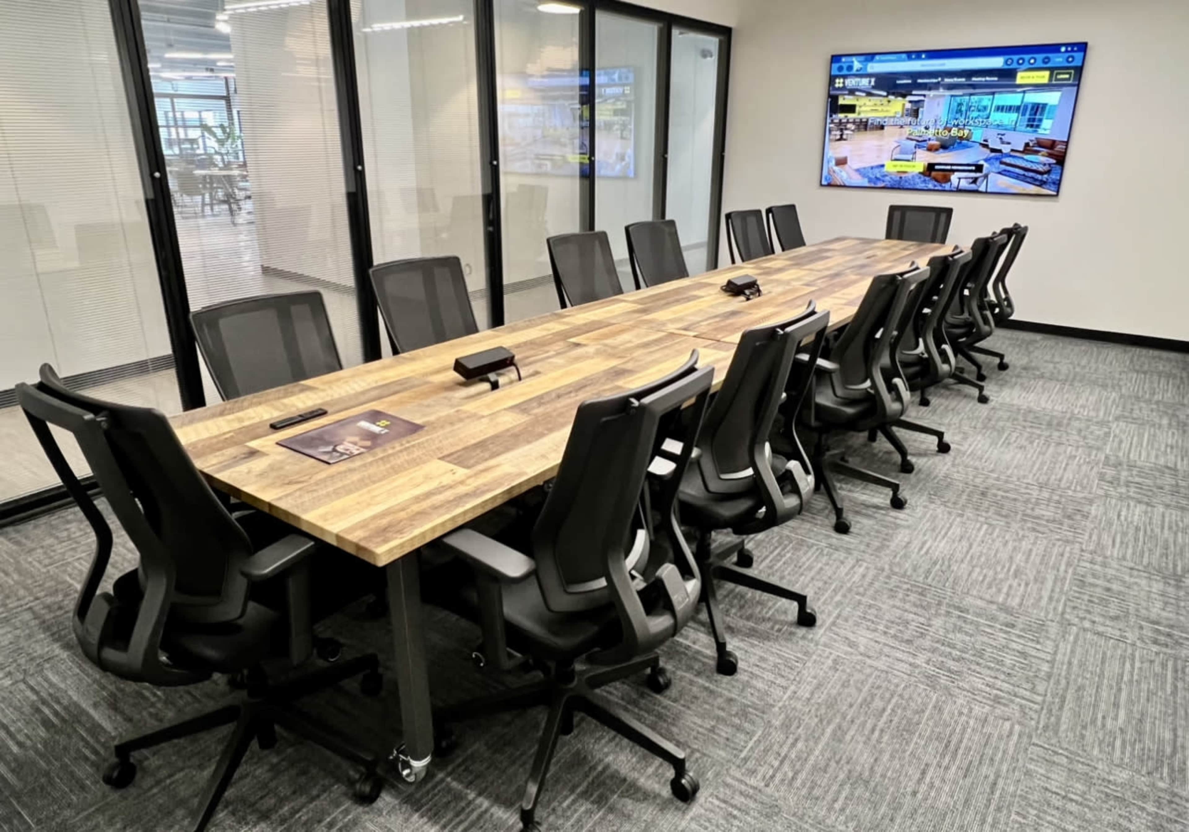 A large conference room features a long wooden table surrounded by ergonomic black chairs, with a wall-mounted screen displaying a digital interface.