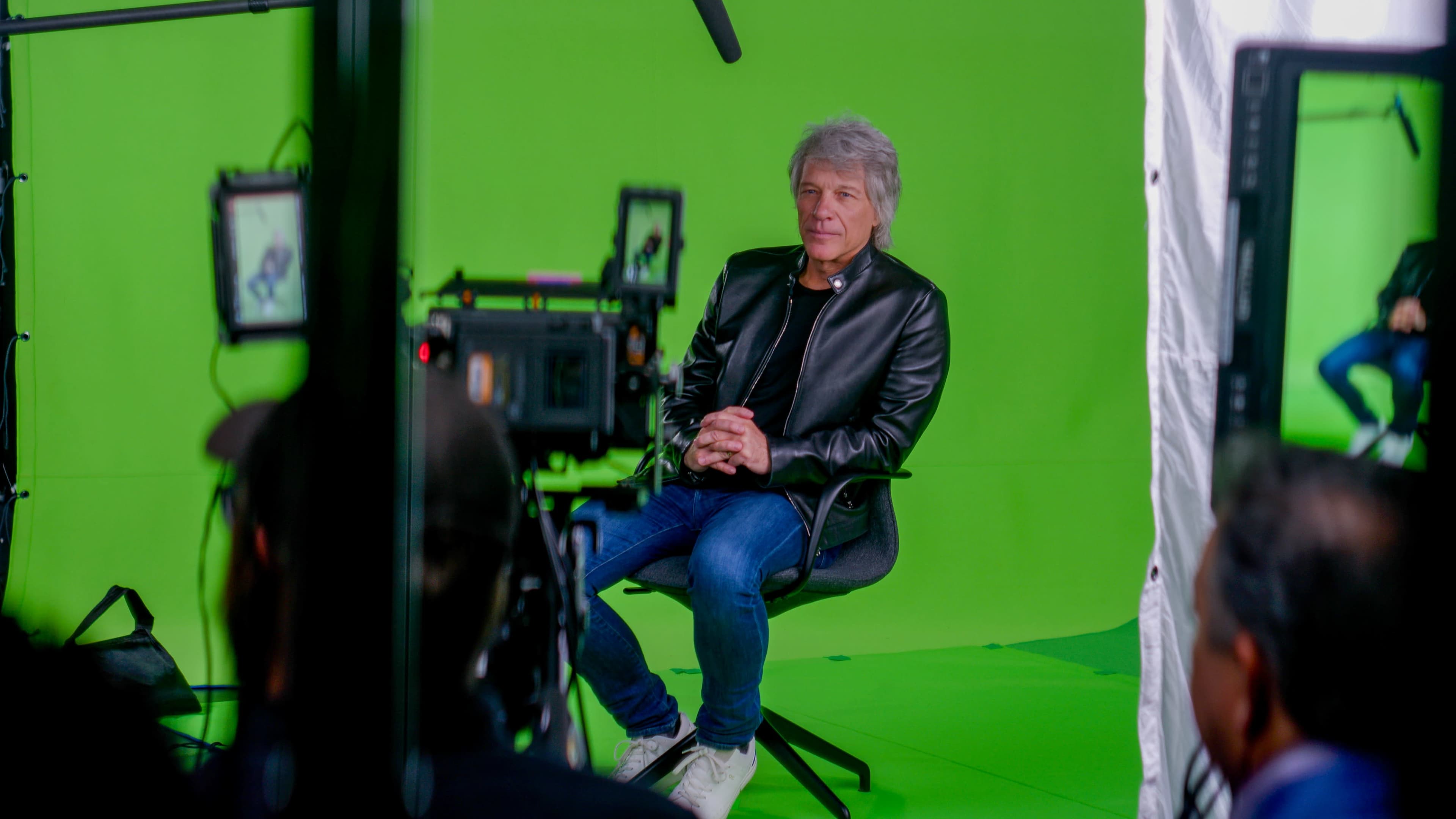 A person with gray hair sits in a chair in front of a green screen, surrounded by camera equipment.