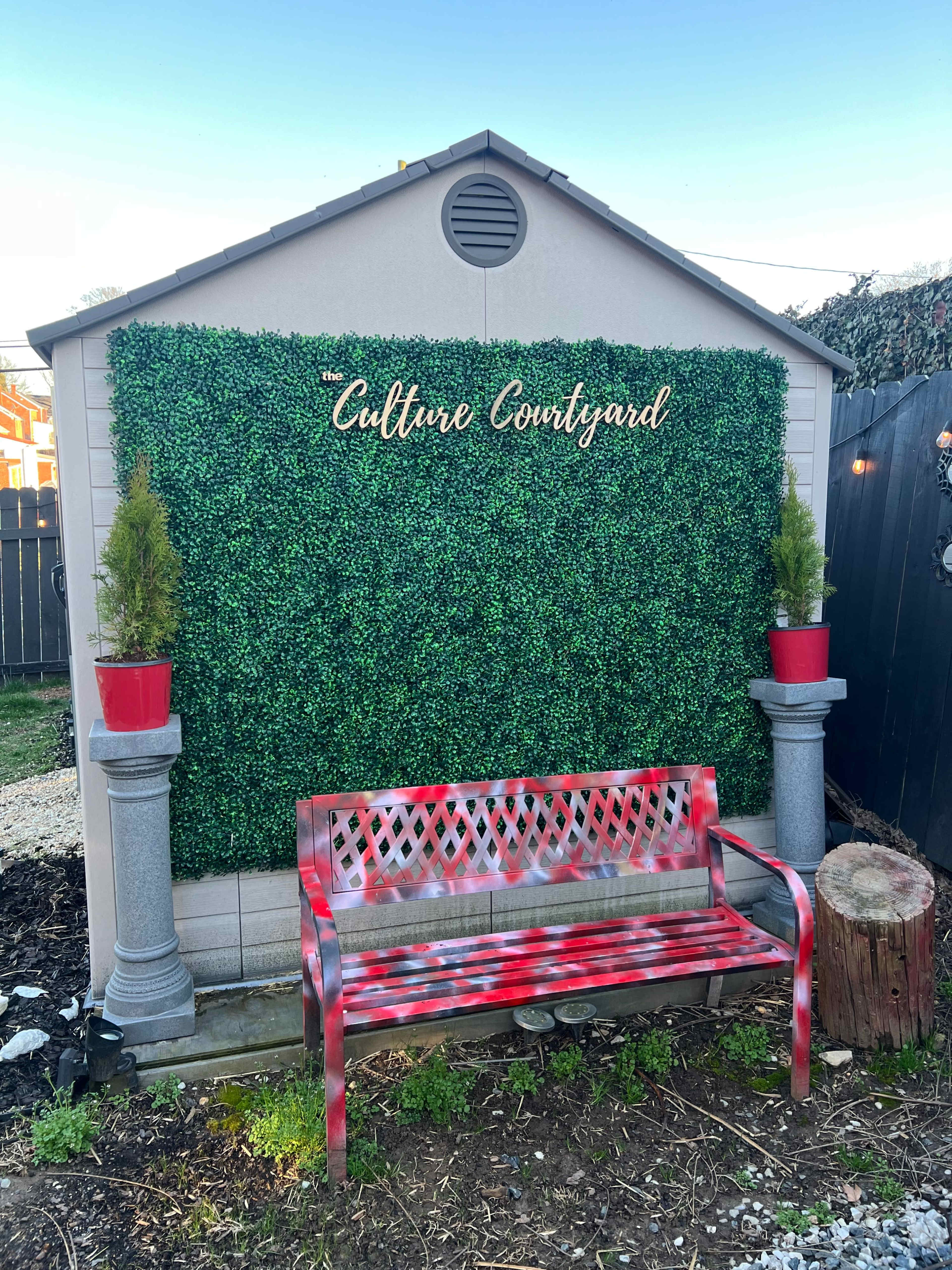 A small shed features a green hedge wall with the text "Culture Courtyard" above a red bench flanked by potted plants.