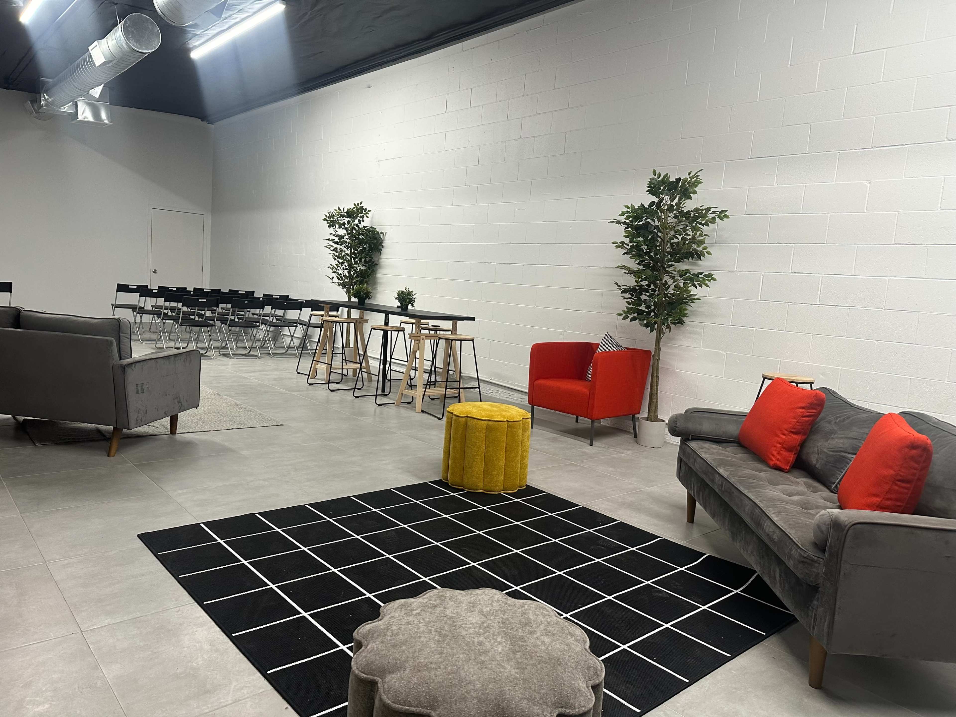 A modern lounge area with gray sofas, a red armchair, a yellow ottoman, and a seating space with black chairs and wooden tables against a white wall.