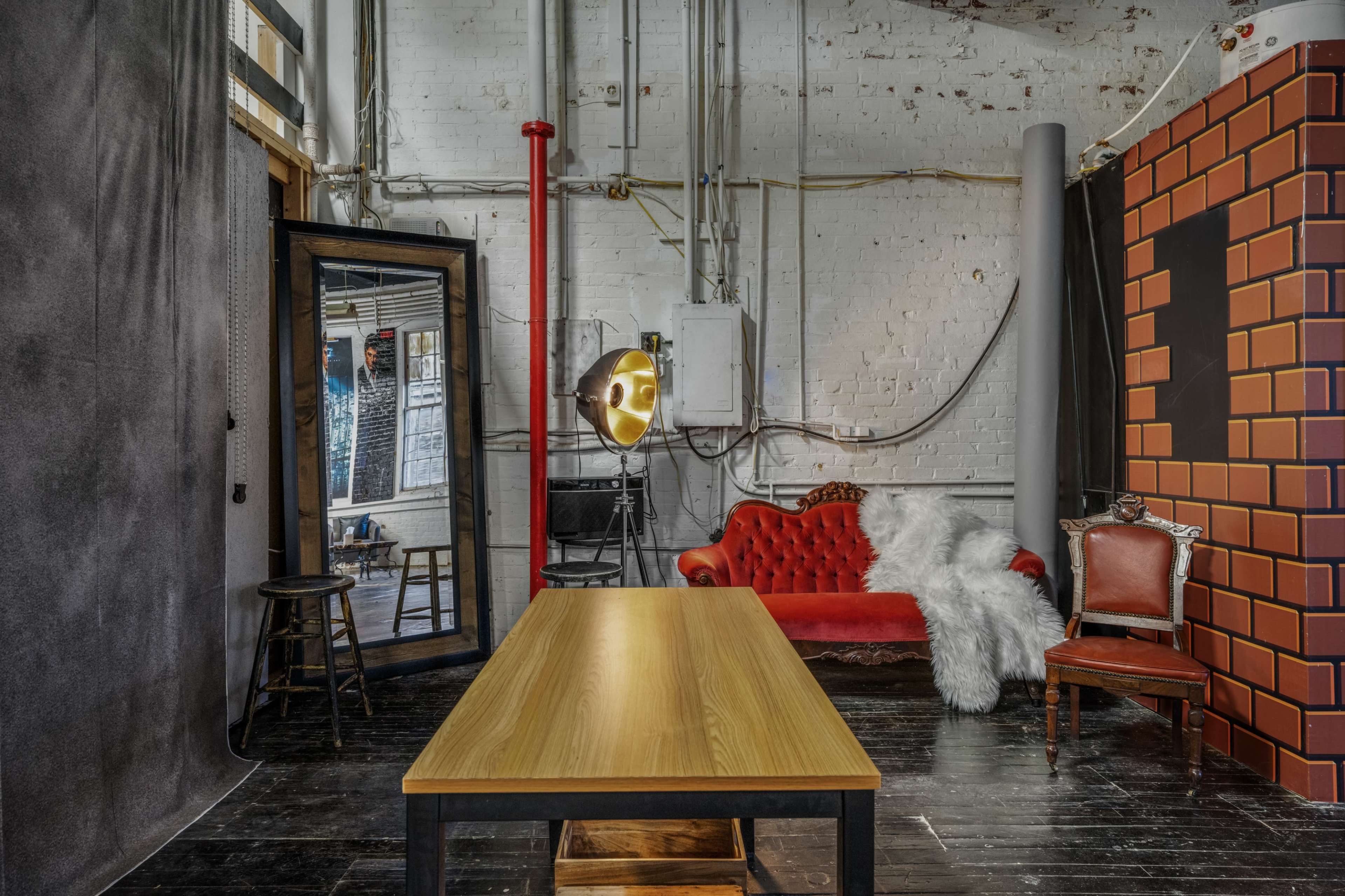A spacious studio interior featuring a red velvet sofa, a wooden table, a large mirror, and industrial-style lighting against a backdrop of exposed brick and pipes.