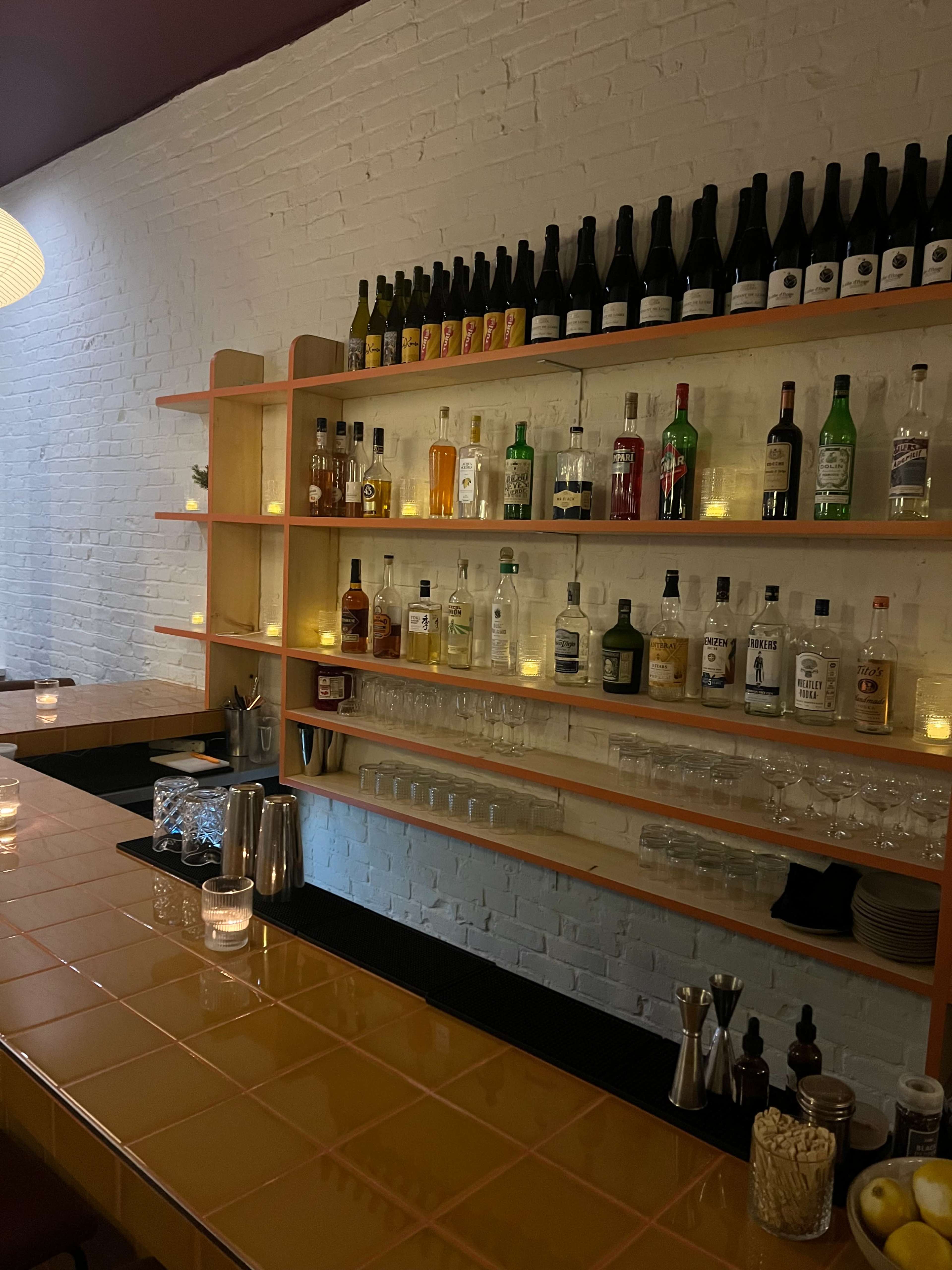 The image shows a neatly arranged shelf of various bottles of alcohol above a bar counter with a tiled surface and warm lighting.