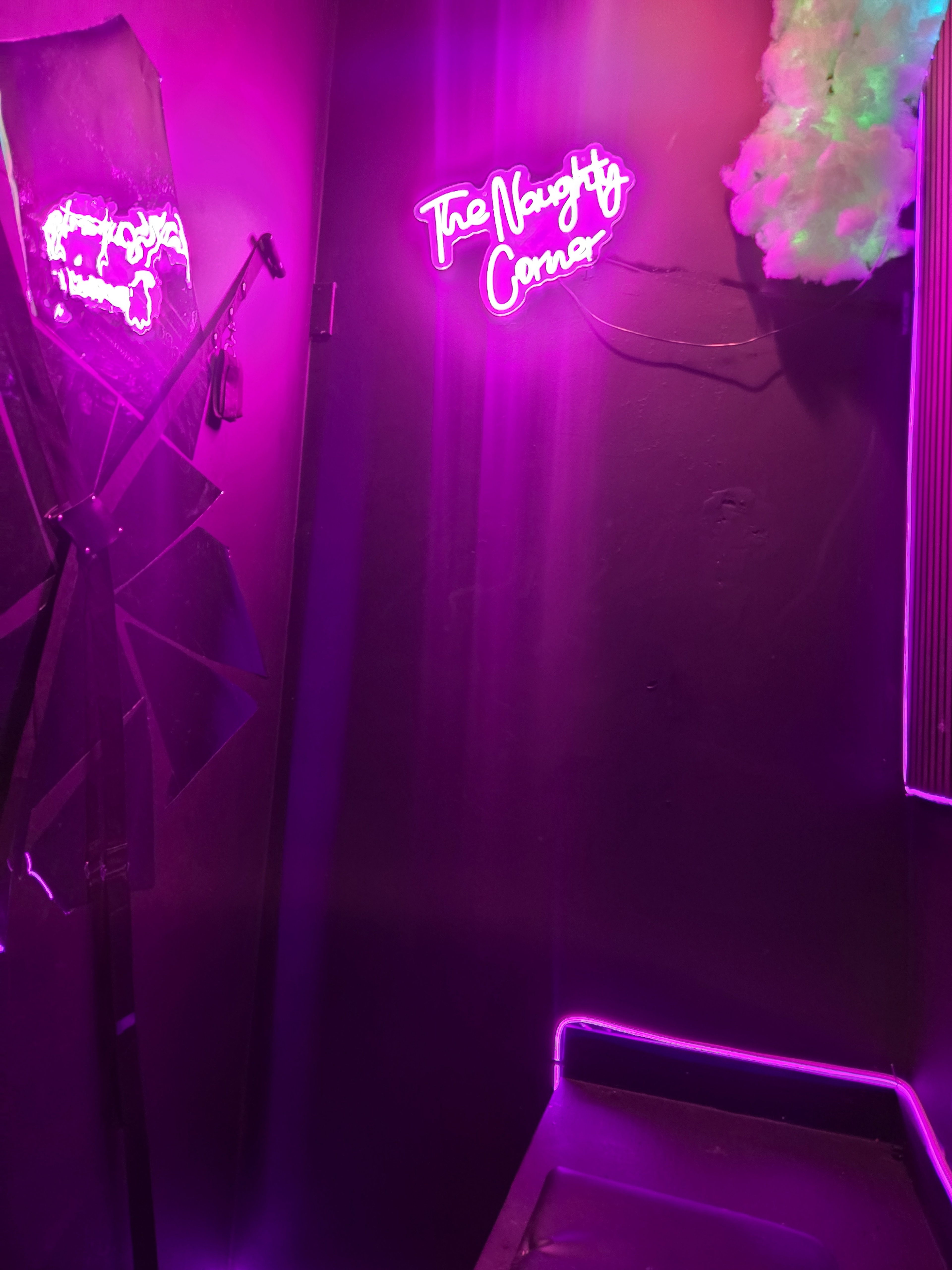 The image shows a dimly lit corner with a neon sign that reads "The Naughty Corner," surrounded by purple lighting and a partially opened umbrella.