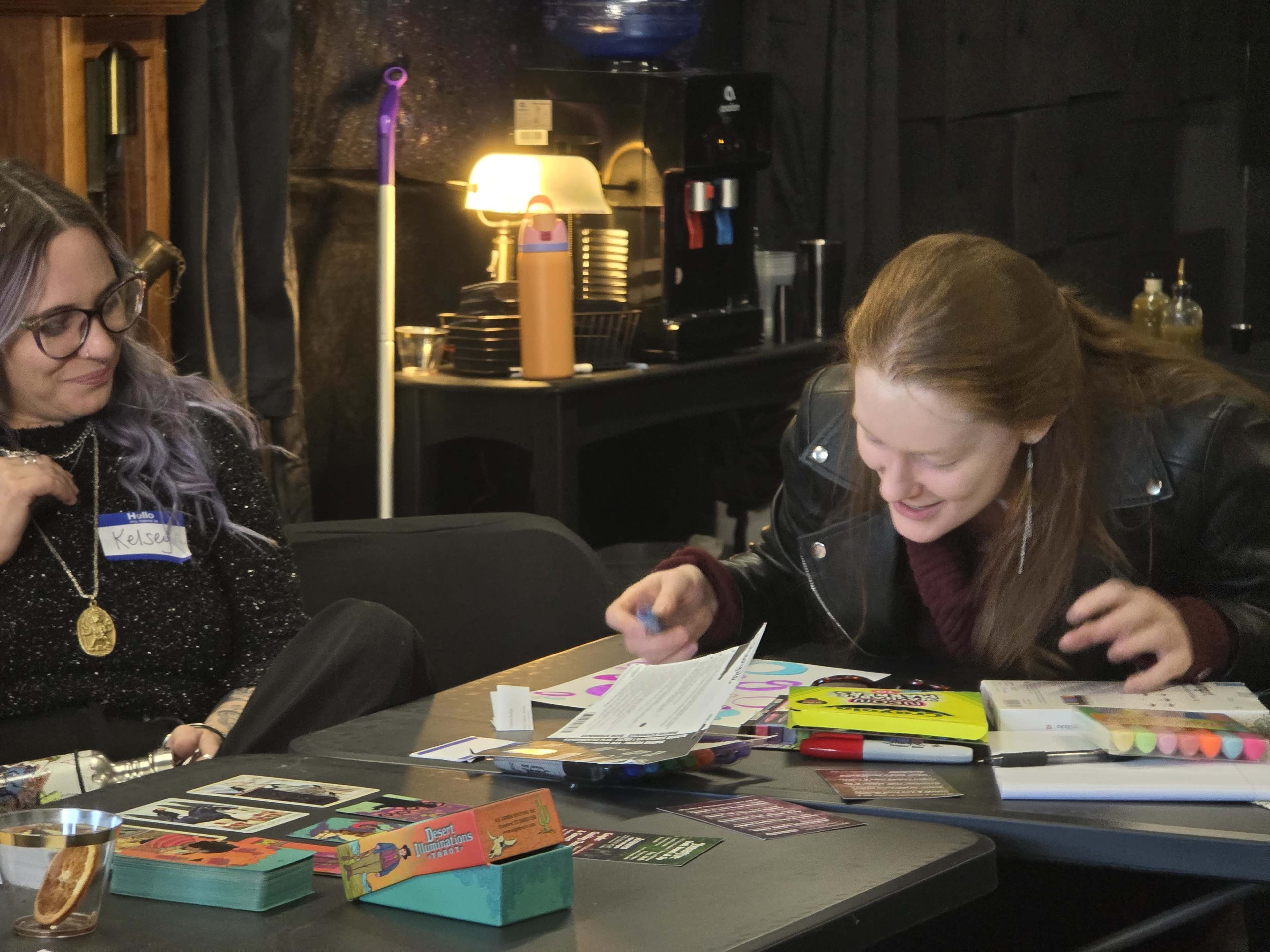 Two women are seated at a table filled with various games and art supplies, with one woman laughing and interacting with the materials while the other observes.