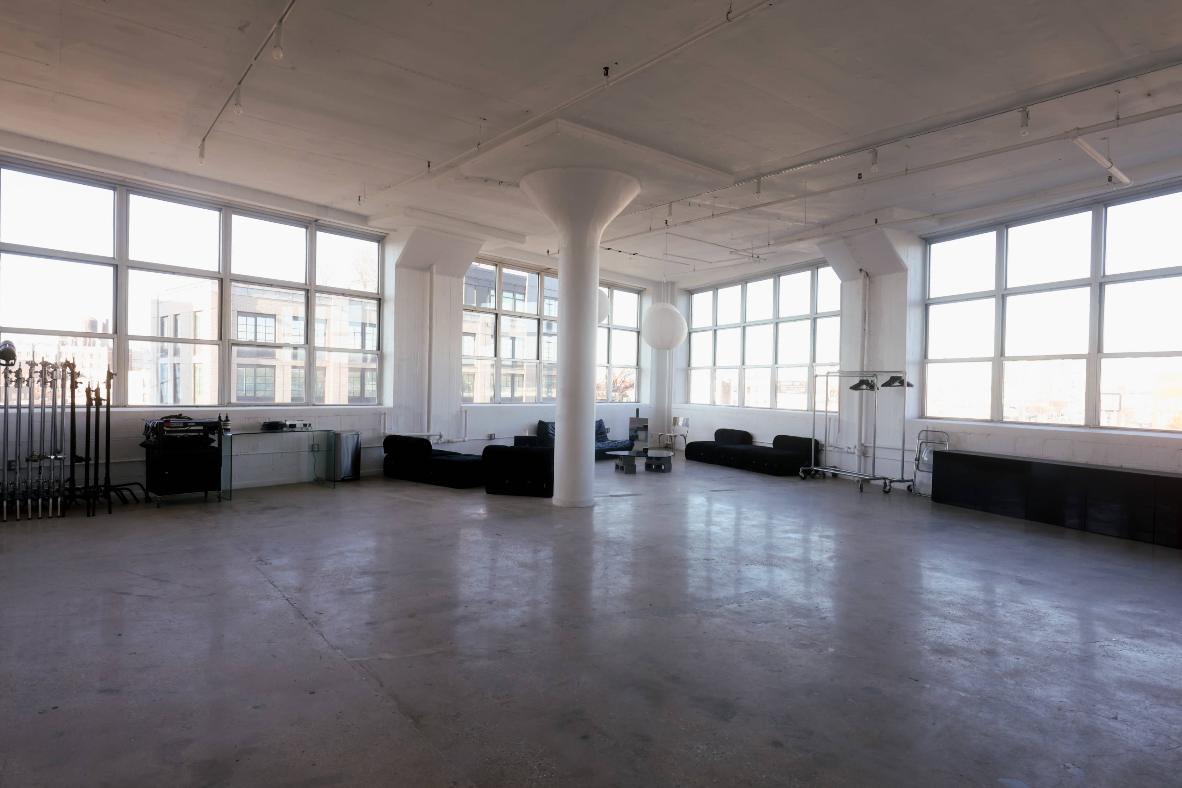 The image shows a spacious, minimalistic room with large windows, concrete floors, and a few black couches arranged around the area.
