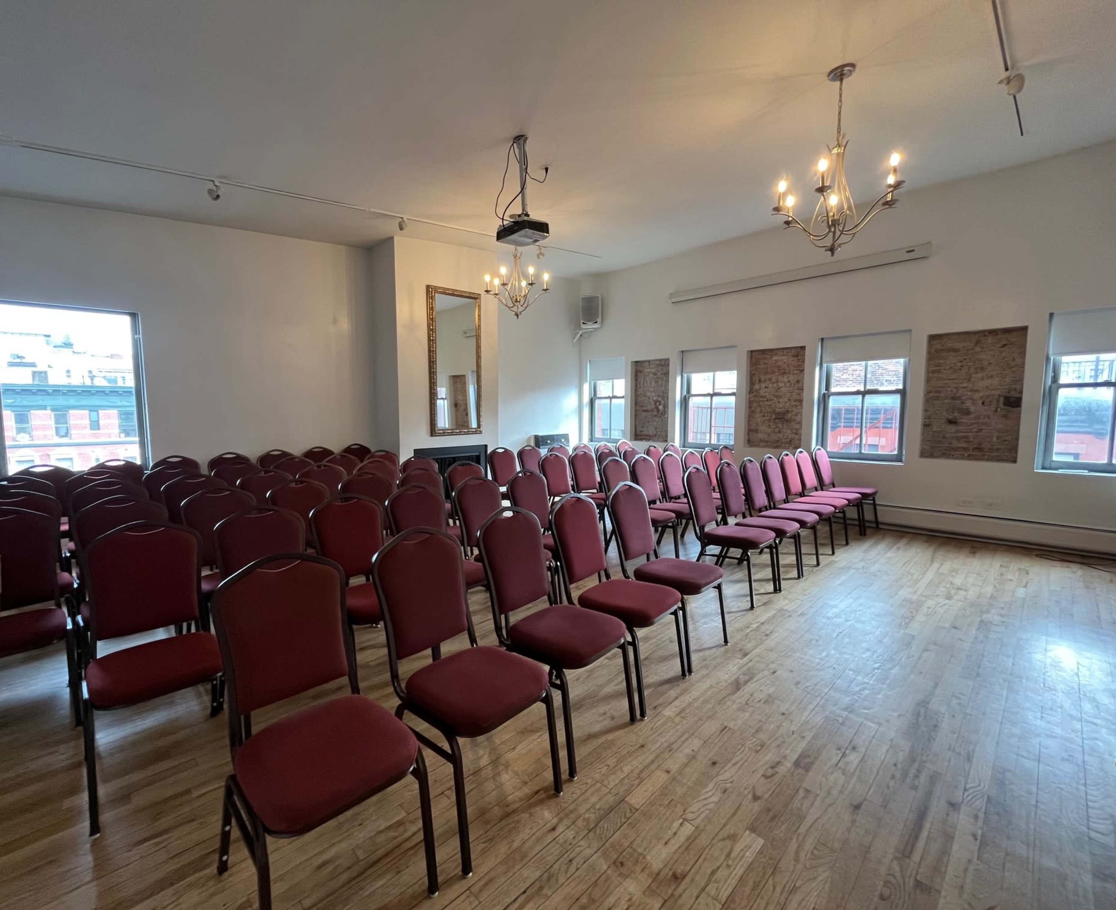 The image shows a spacious room set up with rows of red chairs arranged for an audience, featuring large windows and wooden flooring.