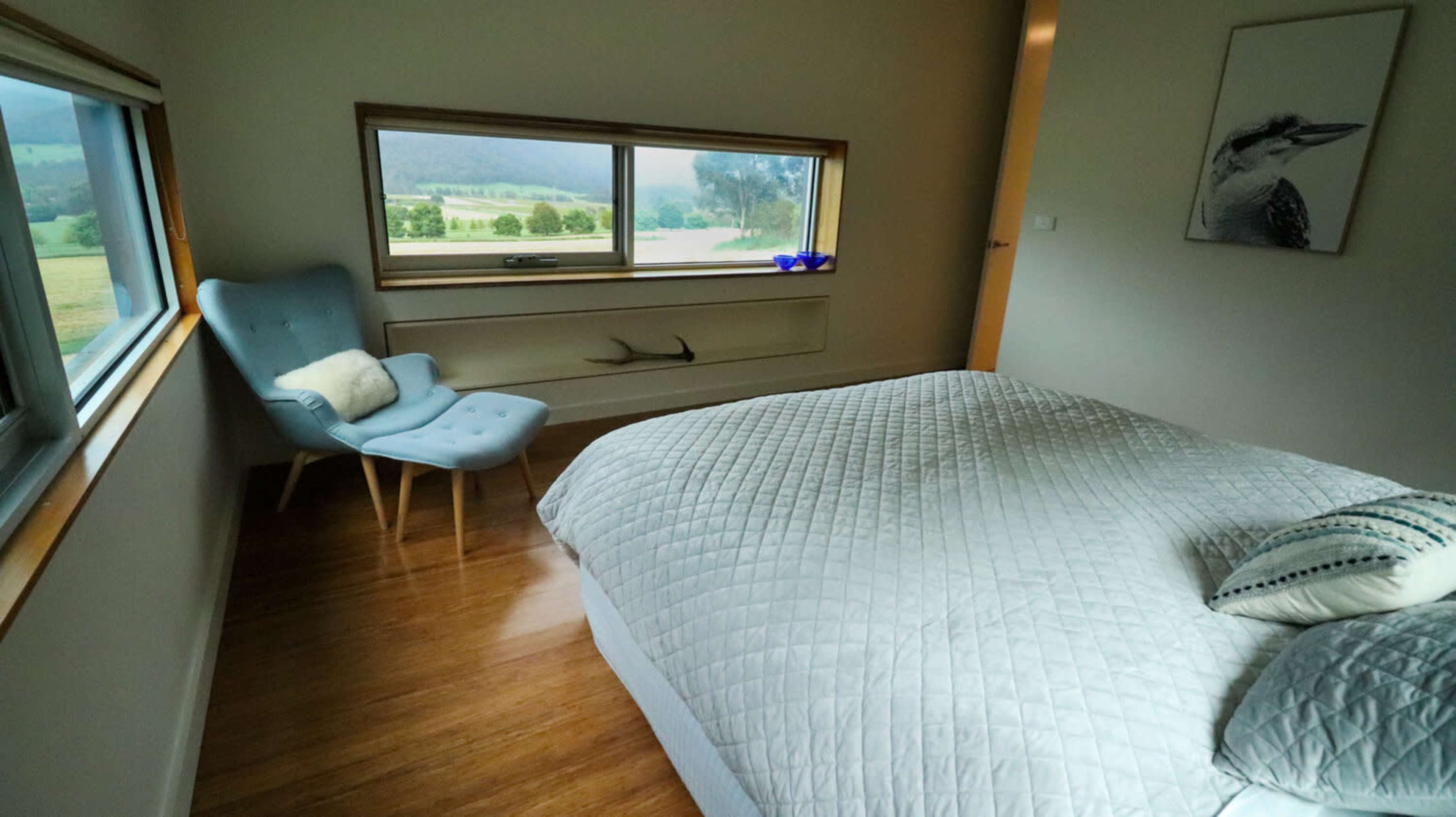A simple bedroom features a bed with a quilted cover, a light blue chair near a window, and a scenic view of green fields and hills outside.