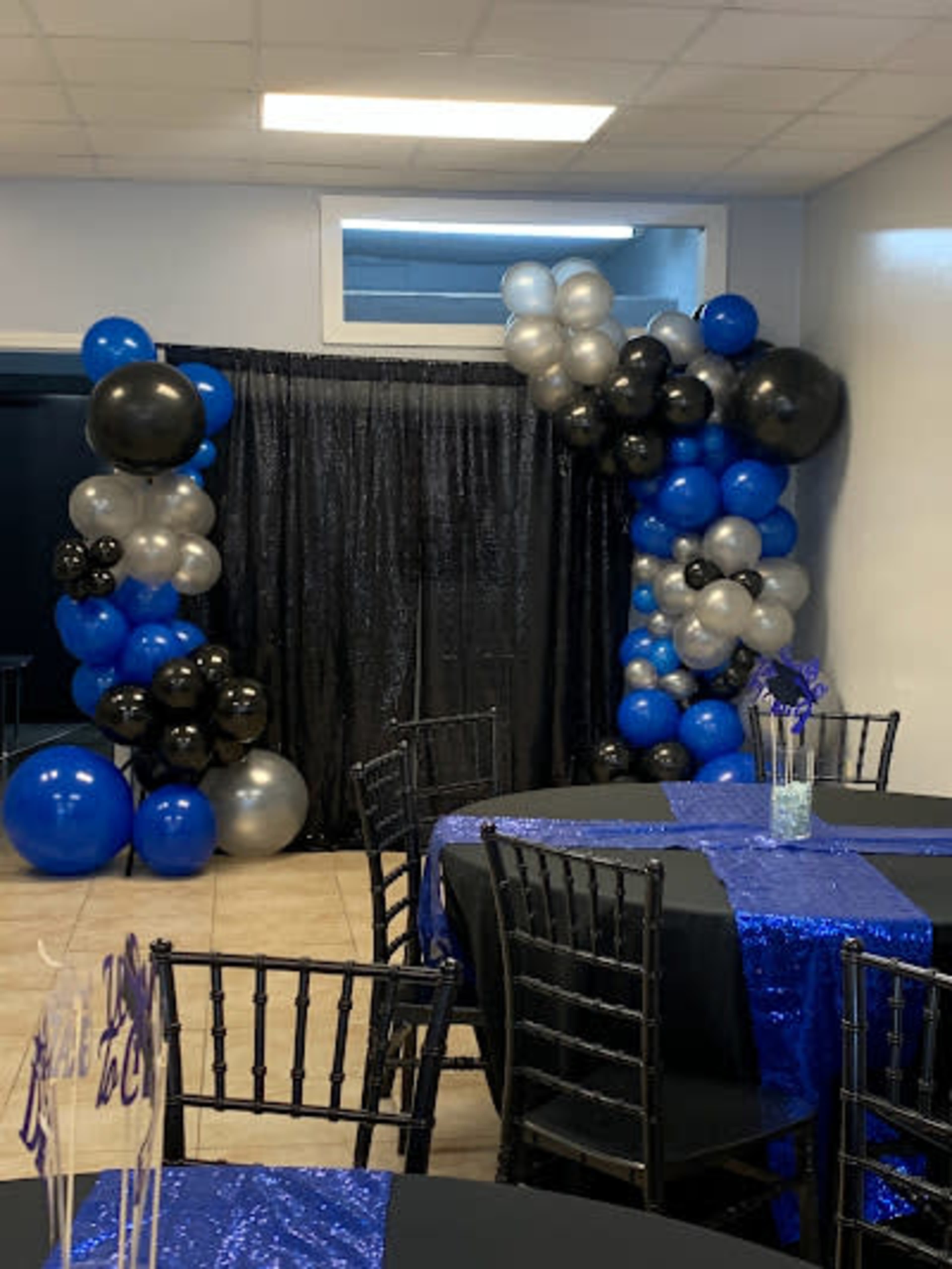 A party venue features a corner decorated with blue, black, and silver balloons beside a black curtain, with tables set in front covered in black and blue sequined tablecloths.