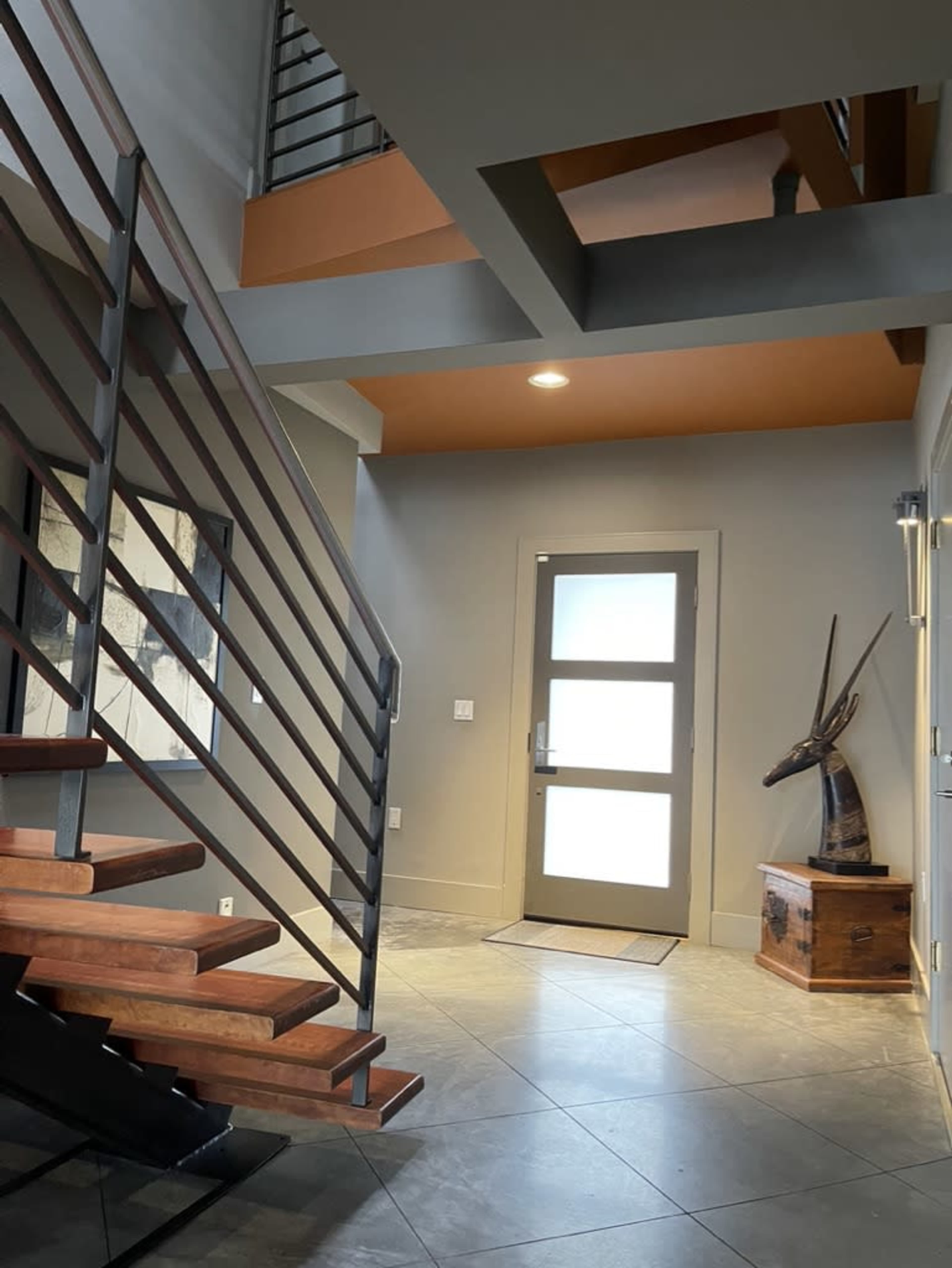 The image shows a modern entryway with wooden stairs leading to an upper level, a glass-paneled door, and a decorative sculpture in the corner.