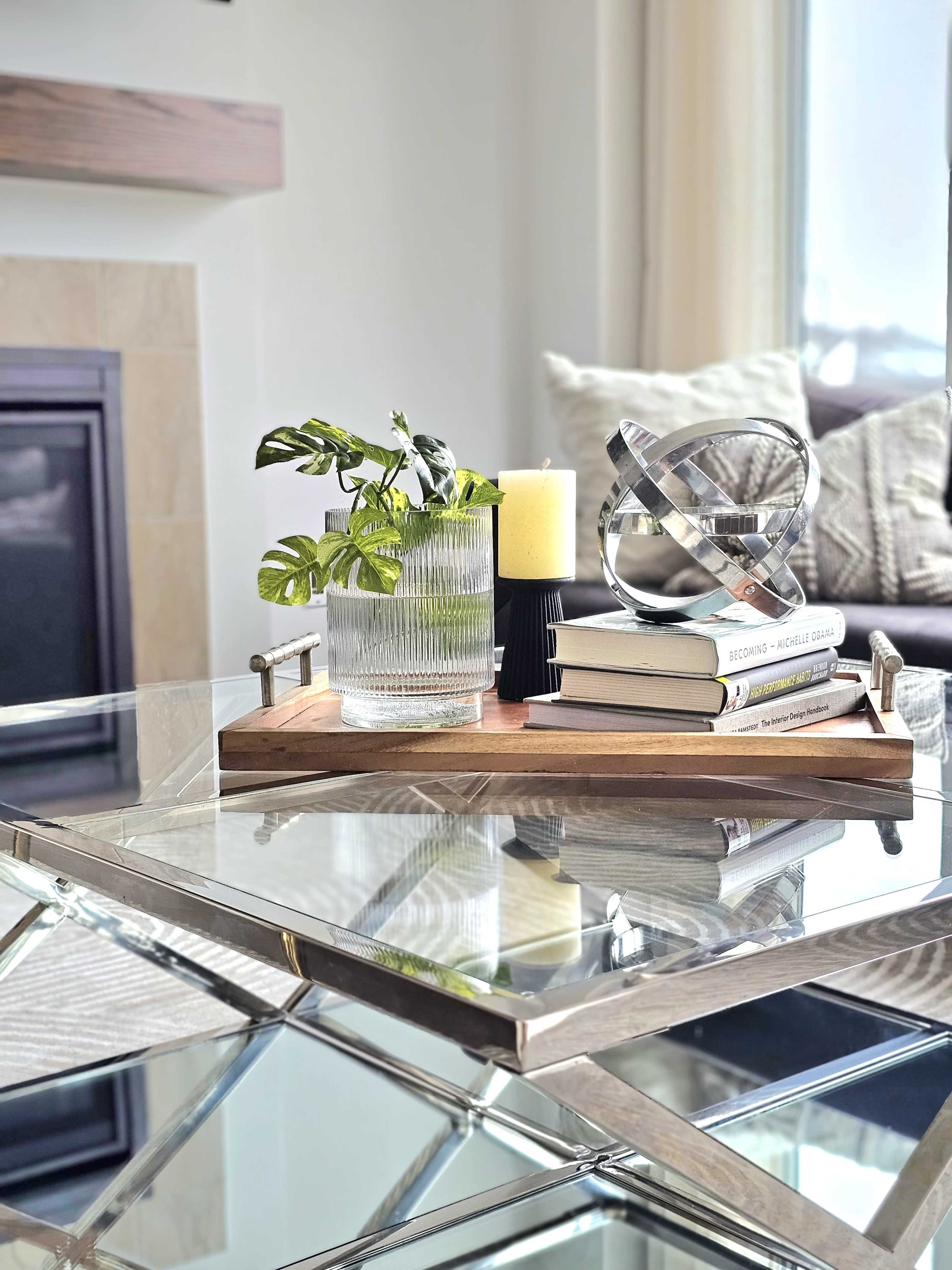 A glass coffee table holds a wooden tray decorated with a plant, a candle, a decorative orb, and several stacked books.
