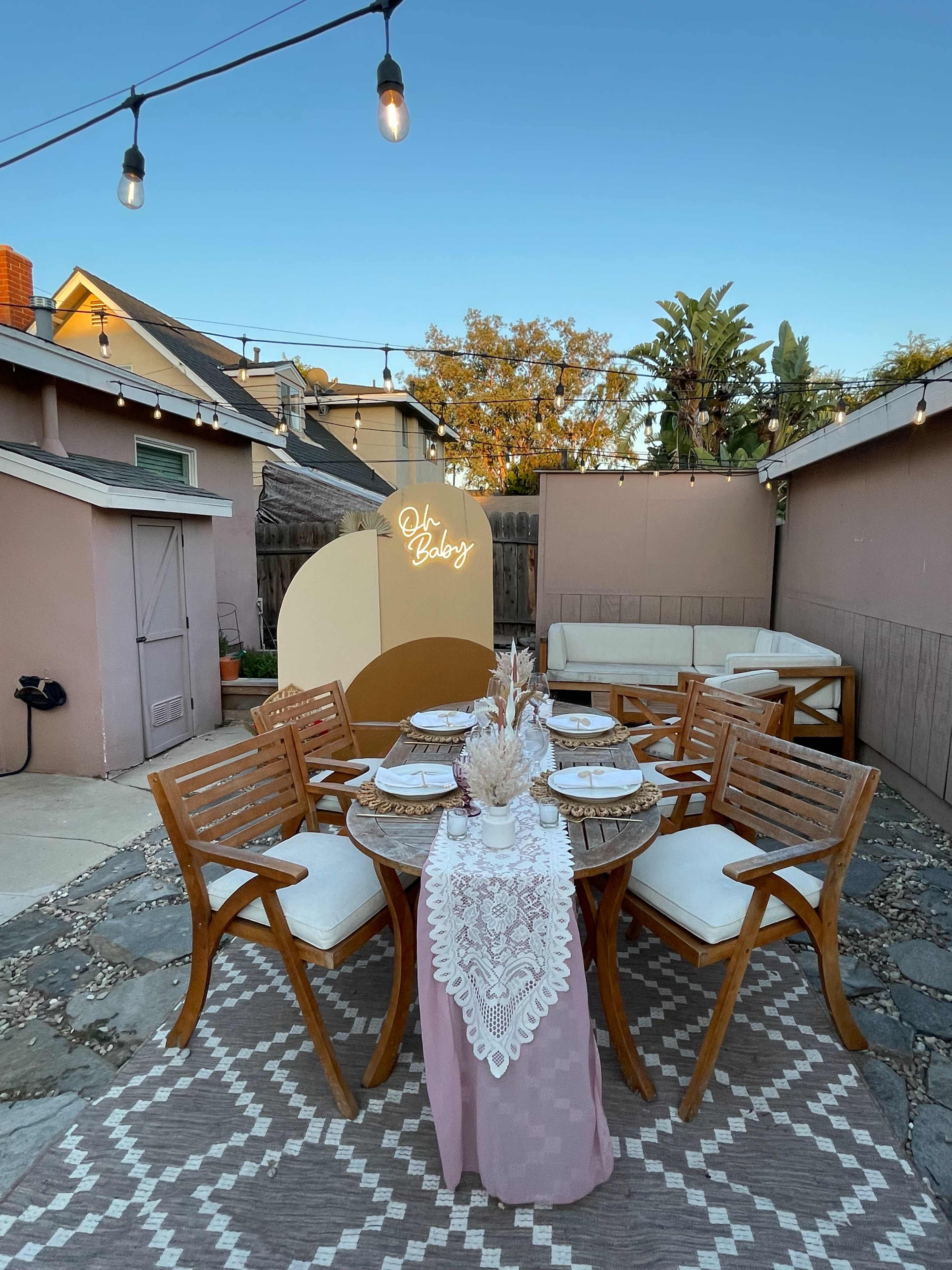 A stylish outdoor dining setup features a round table with lace decor, surrounded by wooden chairs, set against a backdrop of a cozy backyard with string lights and greenery.