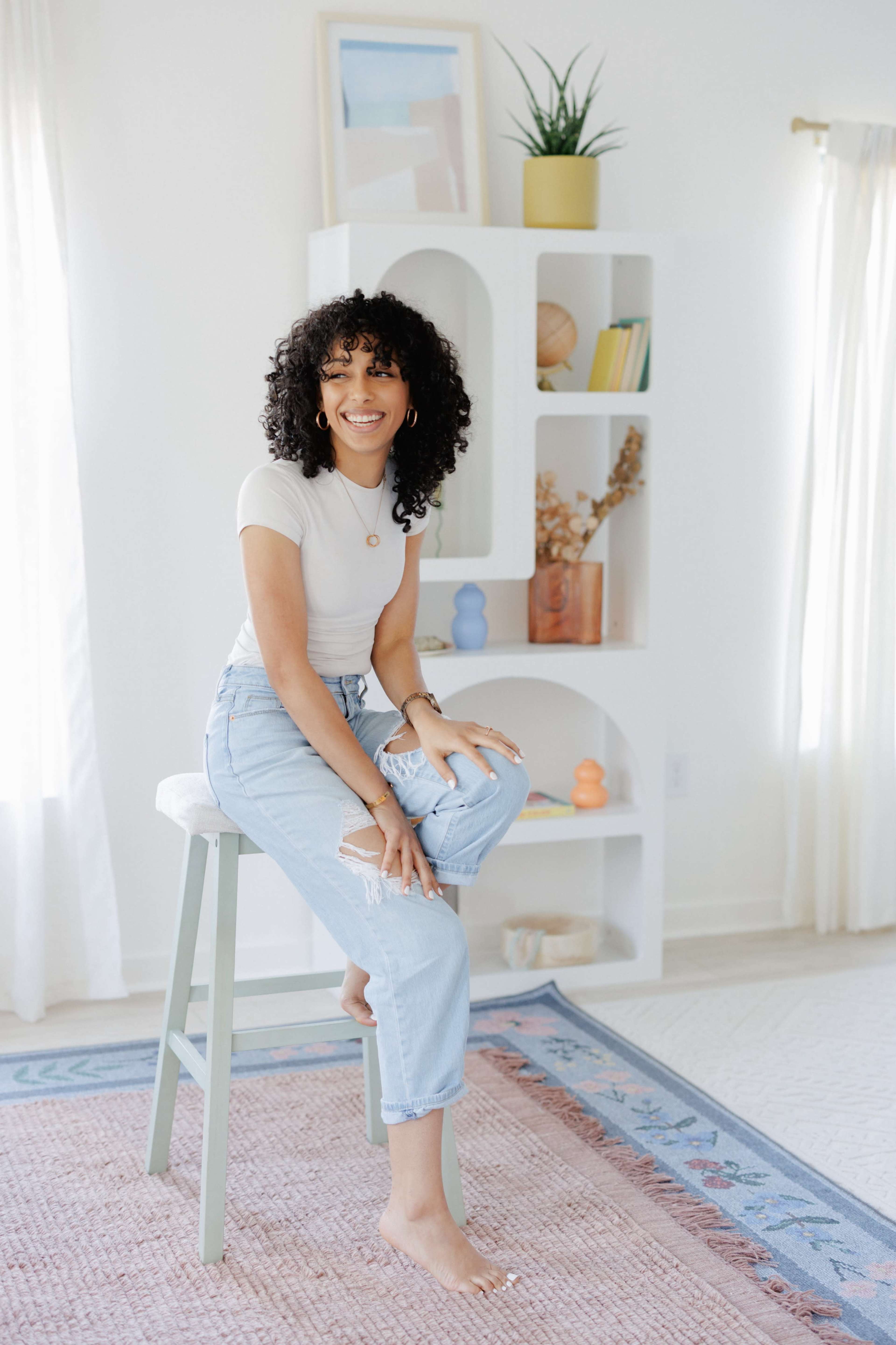 A woman with curly hair is sitting on a stool next to a small white shelf displaying decorative items and plants.